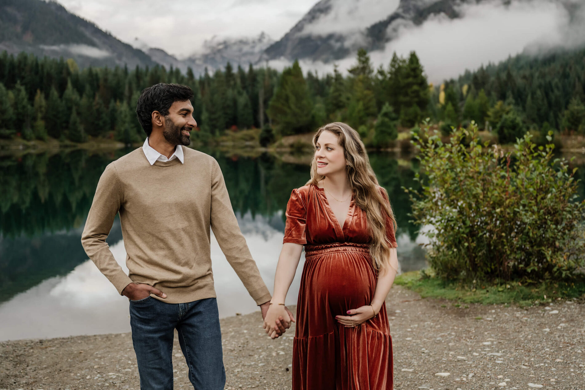 Pregnant woman in a maternity dress holding her husband's hand during a Seattle photoshoot in front of a pond with mountains in the background