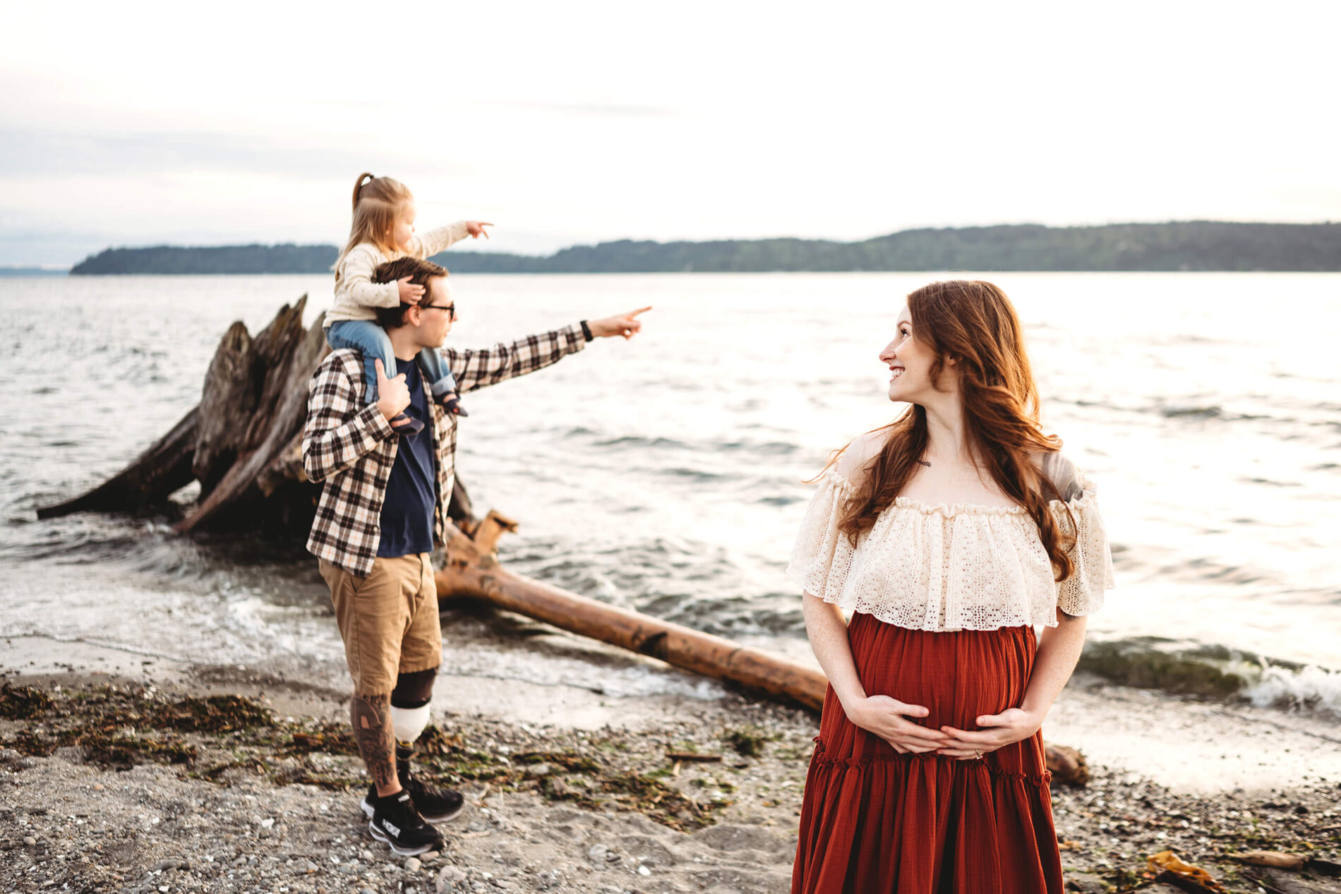 A family maternity photoshoot on a beach in Seattle with a focus on smiling mom, and dad and sibling in the background bonding on a beach