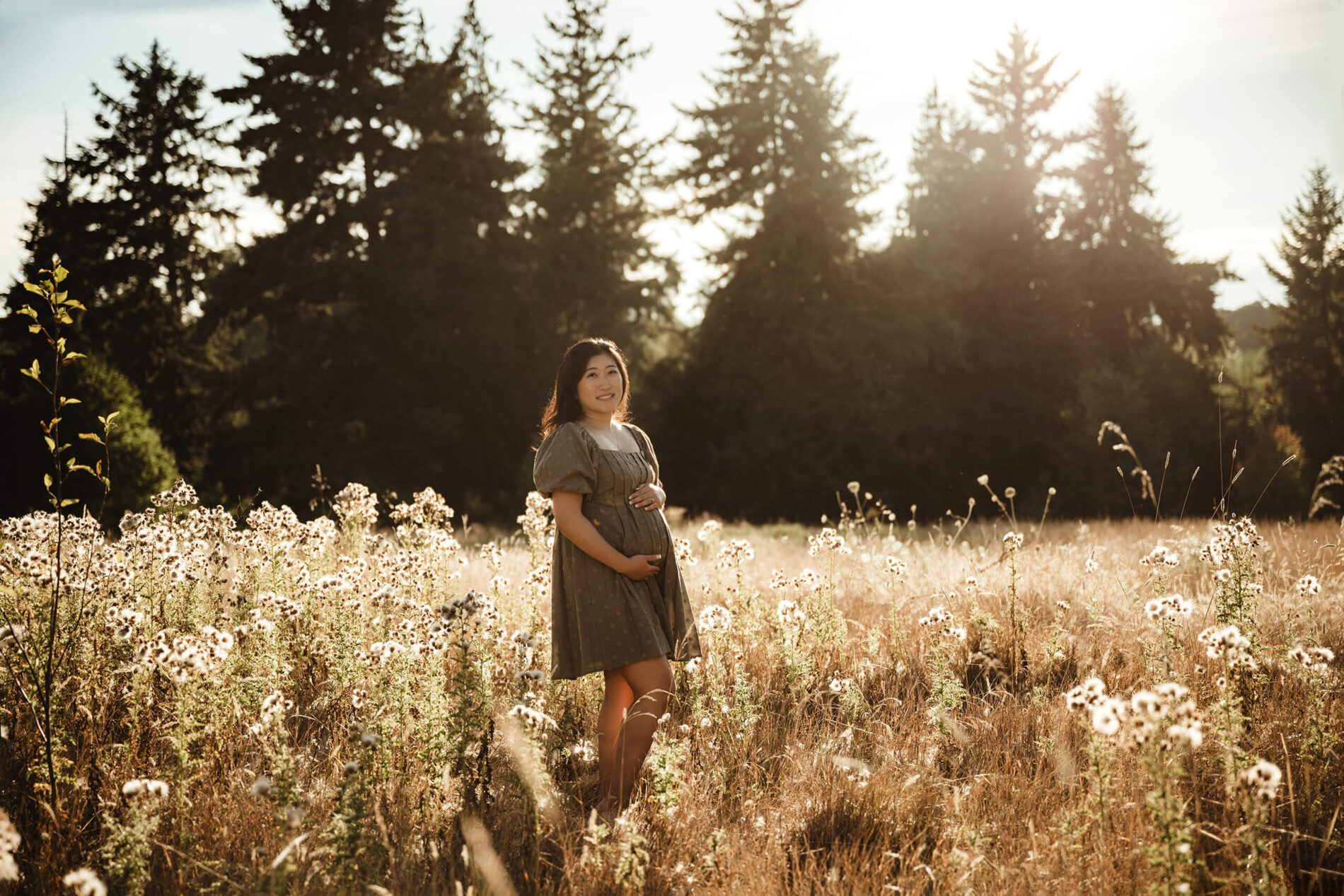 A pregnant woman looking directly into the camera, with her hands wrapped around her belly, in a grass field with wildflowers during maternity photo session in Seattle