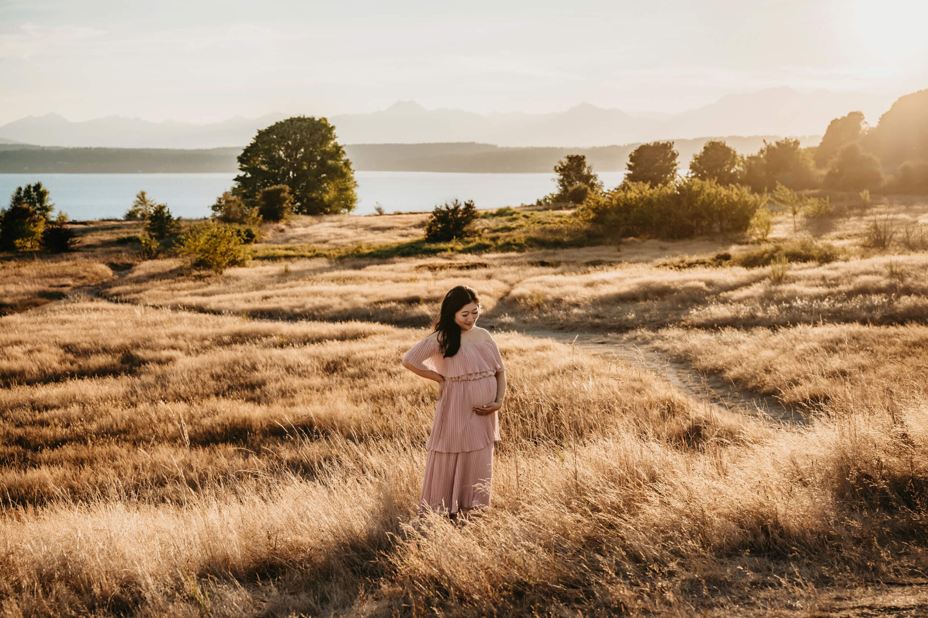 A young pregnant woman in a dress during a photoshoot, standing in a field with tall grass with Puget Sound and Olympic Mountains in the background during sunset