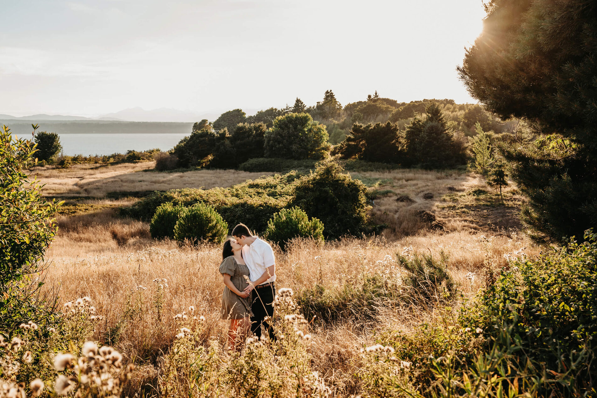 A young pregnant couple kissing during a photoshoot in a field with tall grass with Puget Sound and Olympic Mountains in the background