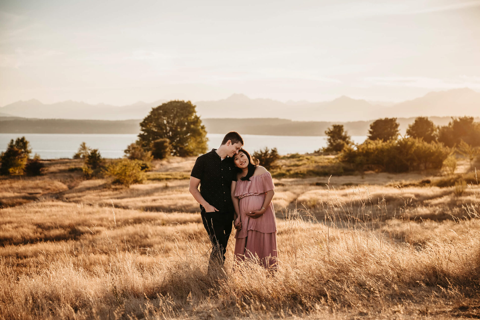 Pregnant couple posed in a beautiful grass field with Puget Sound in the background in Seattle Discovery Park