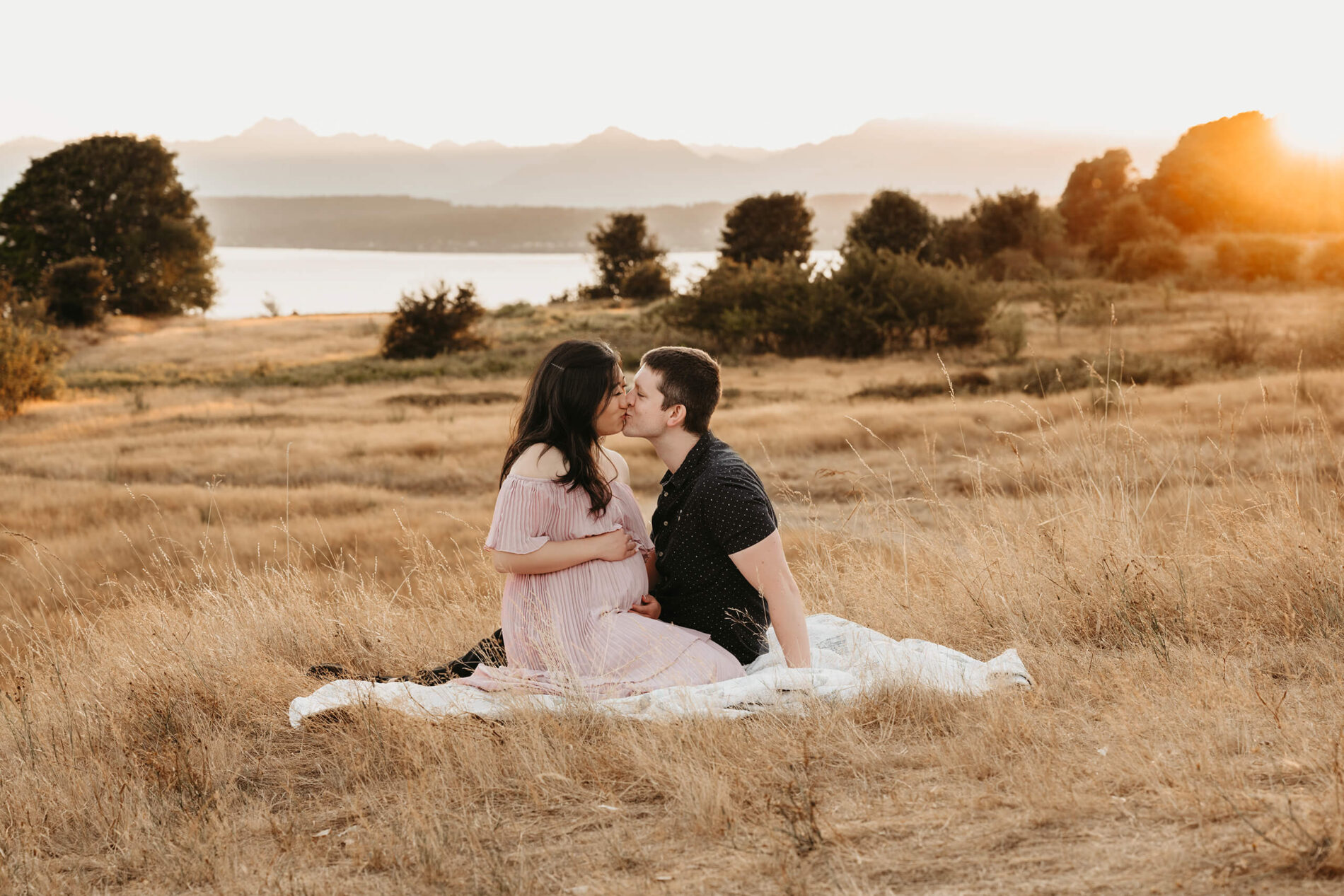 A pregnant couple sitting in an open field in Seattle area, kissing during a maternity photoshoot