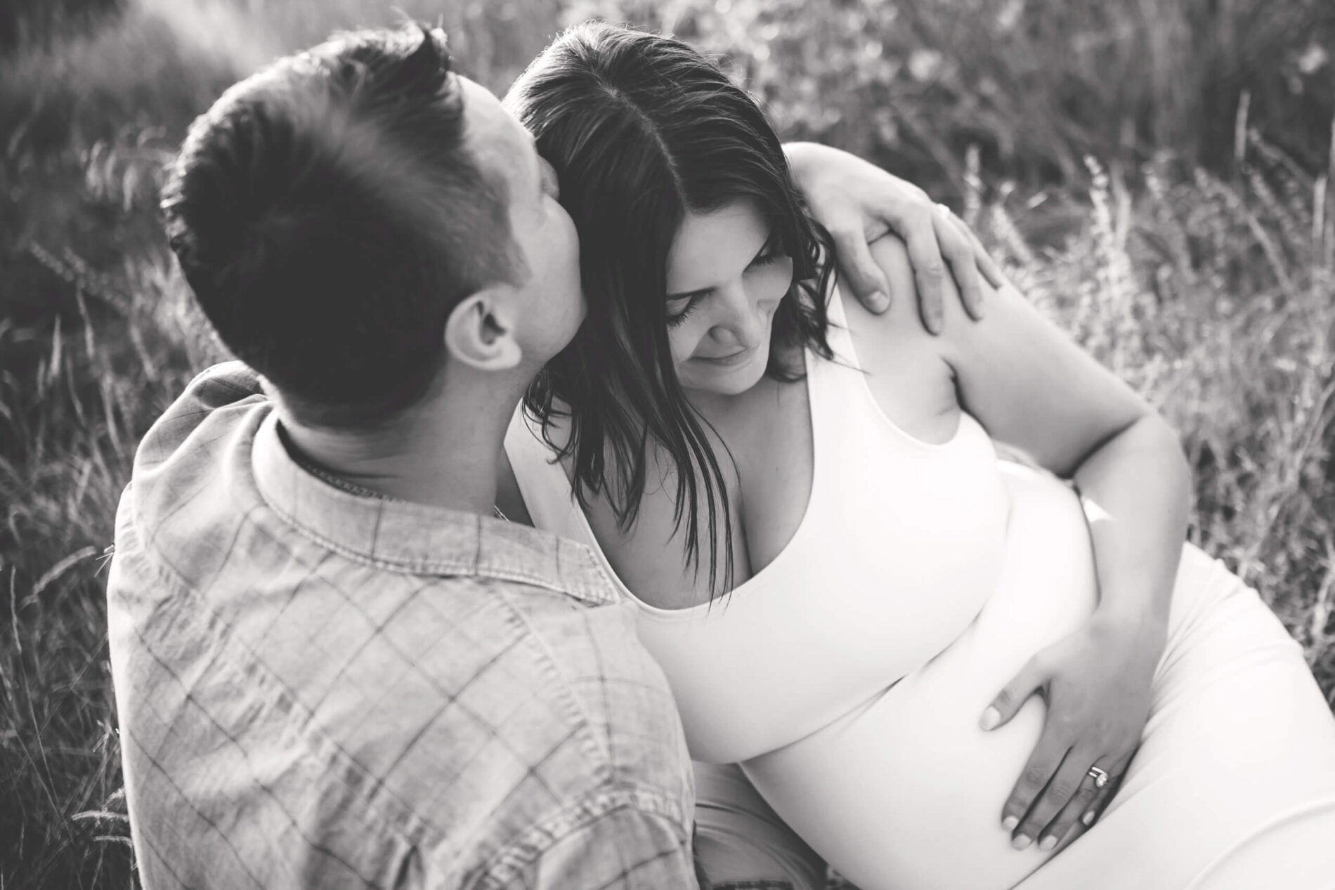 Black and white photo of a couple sitting in a field, hugging, during a maternity photoshoot