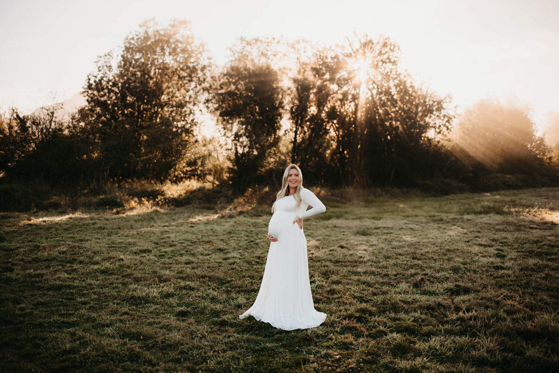 A pregnant woman in a white dress in an open field during sunrise maternity photo session in Seattle area