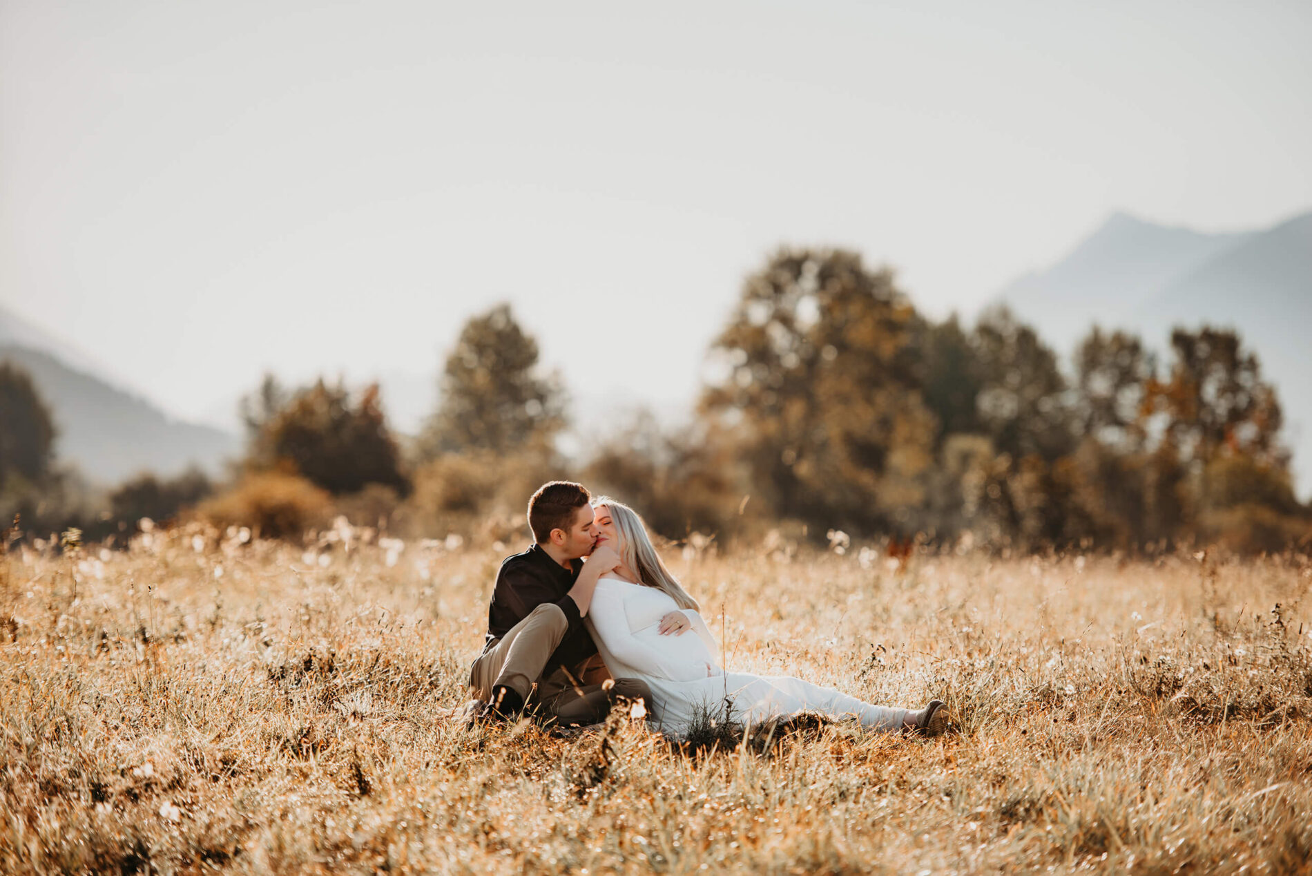 A pregnant couple sitting and kissing in an open field with Mt. Si in the background
