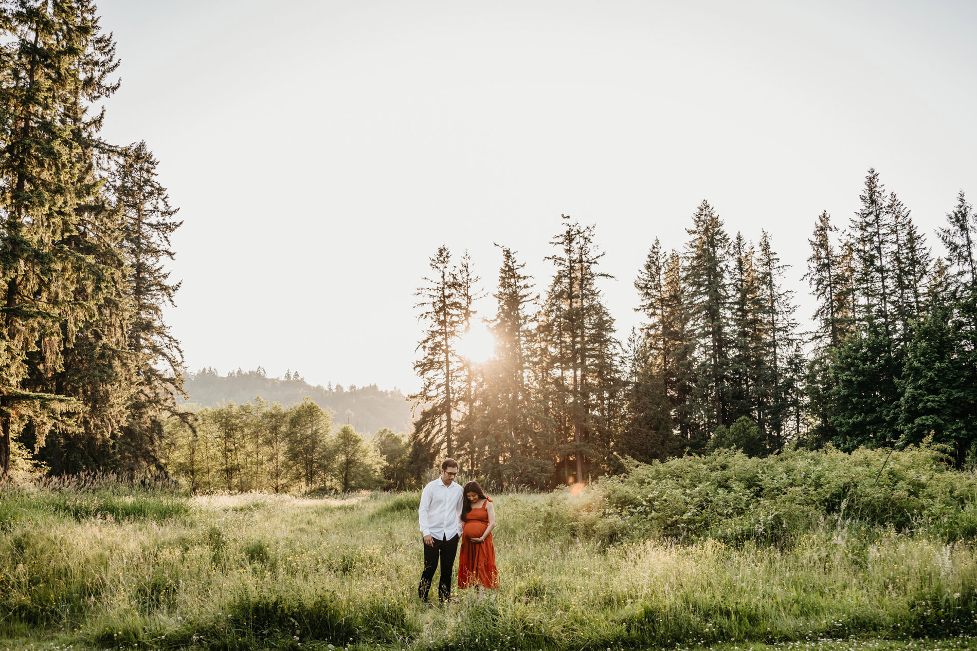 A couple posed in a field of tall green grass, backlit with beautiful warm light during a maternity session in Redmond WA