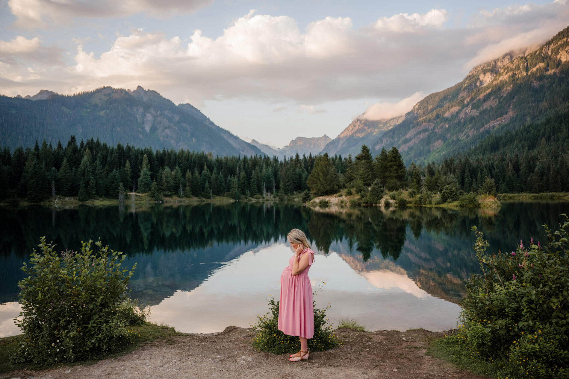 A pregnant woman in a pink dress posed in front of the pond, Cascades mountains in the background, crisply reflected in the pond