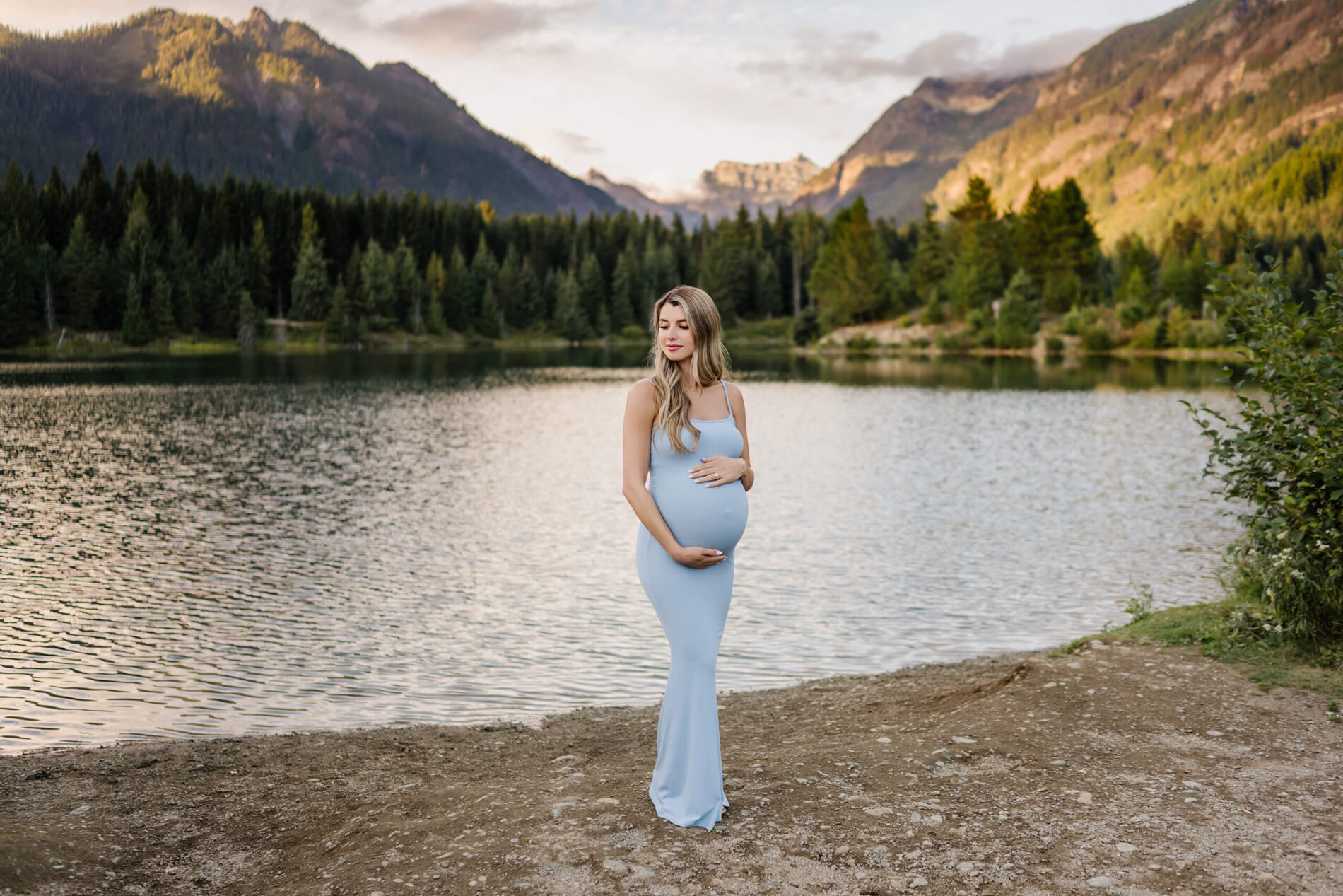 A pregnant woman in a light blue dress in front of a pond with Cascades in the background during a maternity photo session