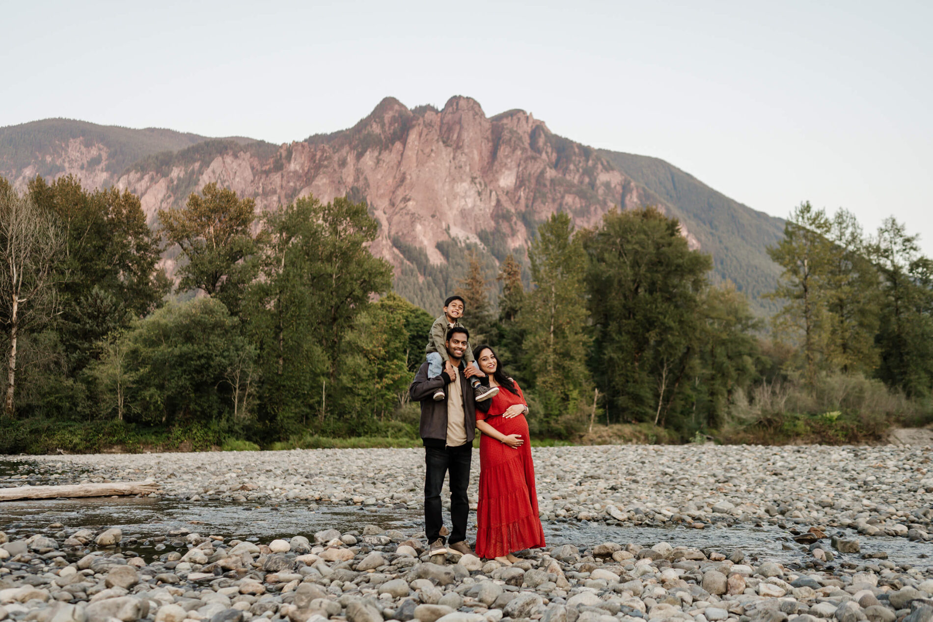 Pregnant woman in a red dress leaning on her husband on a riverbed with Mt. Si in the background