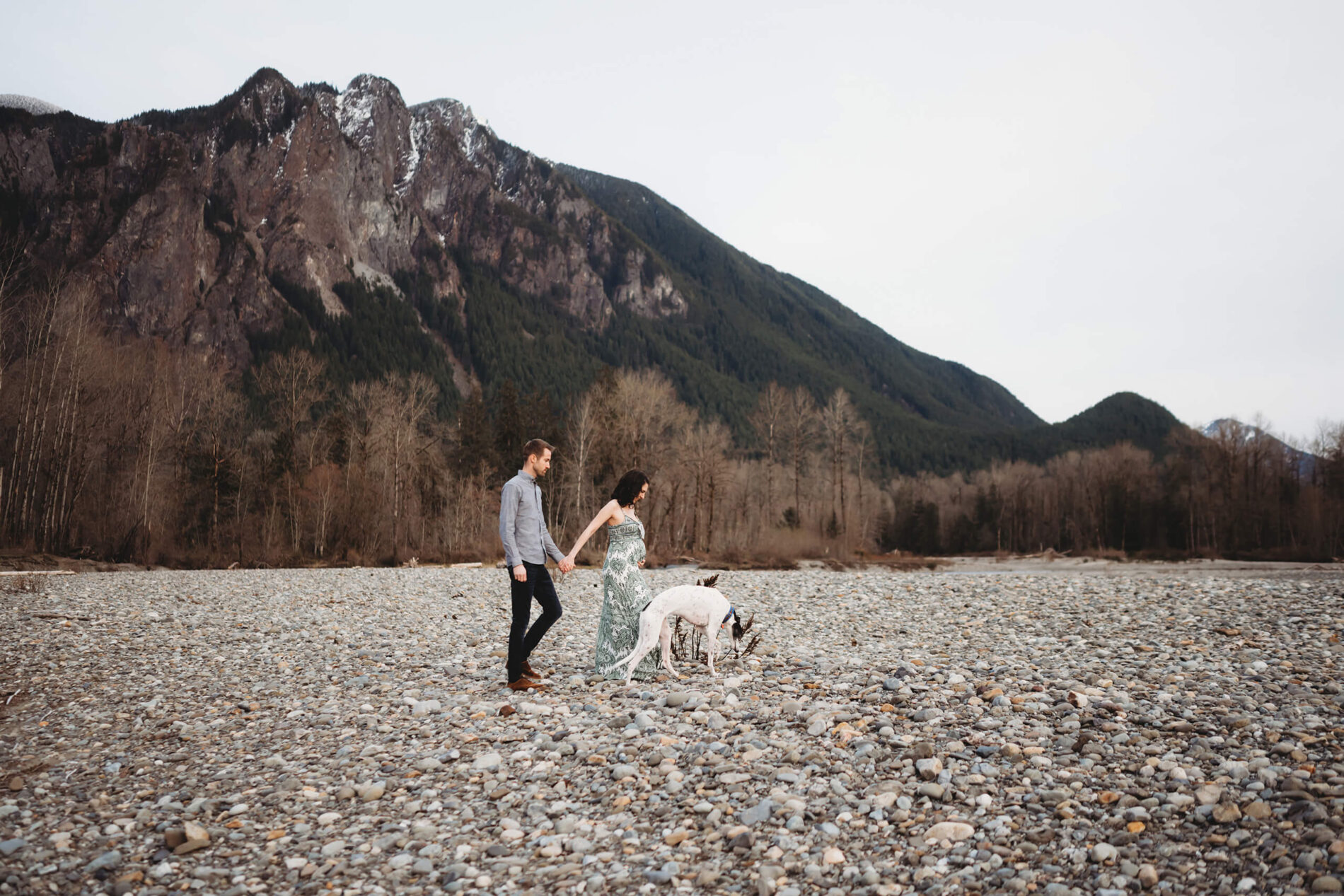 A young couple with their dog walking on a riverbed during a maternity photoshoot with PNW views of breathtaking mountains in the background