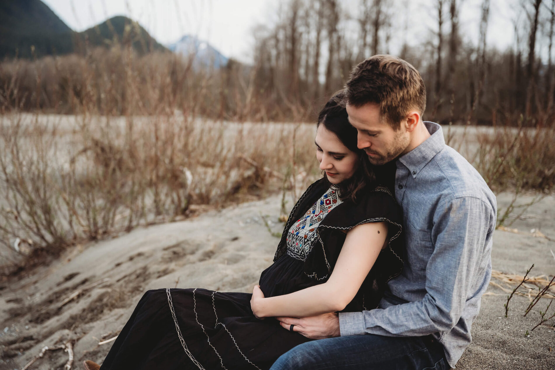 A young couple sitting on a rock during a maternity photoshoot with PNW views in the background