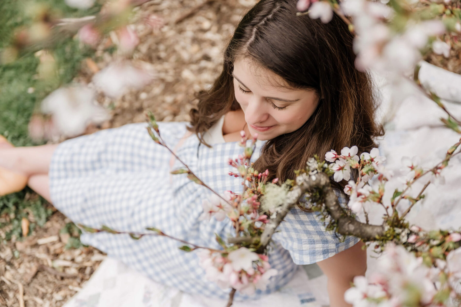 A pregnant woman sitting under a cherry blossom tree, smiling during a photo session at WA Park Arboretum