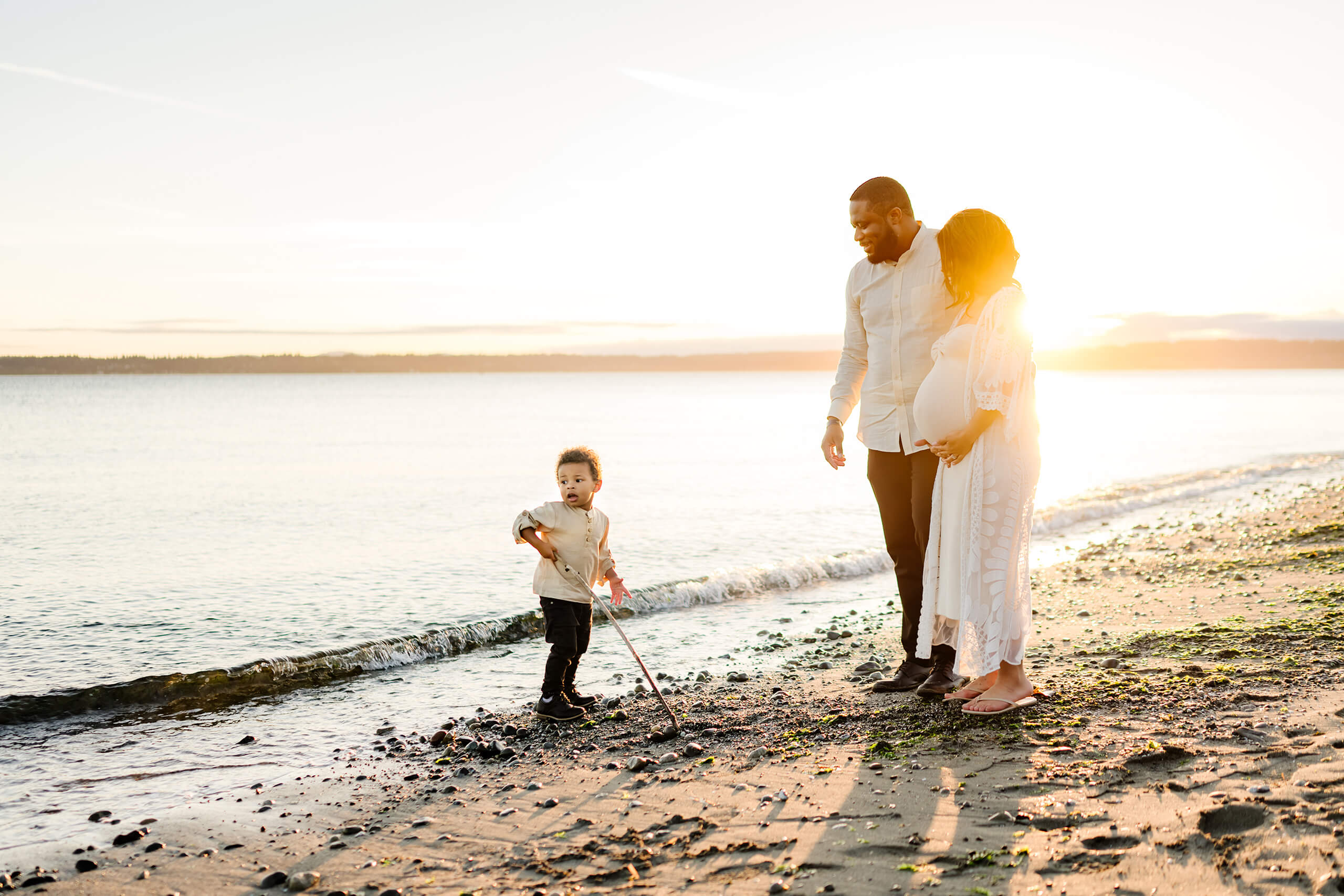 Beach maternity photoshoot at Seattle Discovery Park. Pregnant couple watching their toddler son play on the beach during sunset.