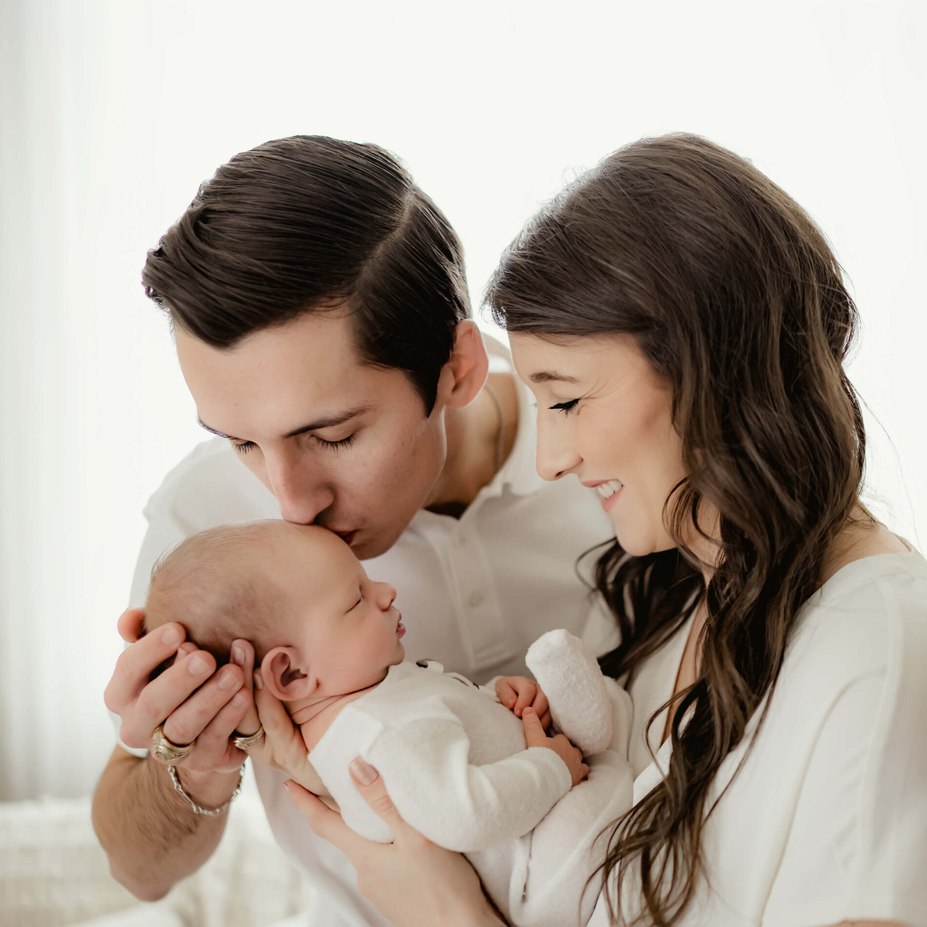 Unique newborn photography with parents. Mom holding her son on her chest, with dad gently kissing his son on his head.