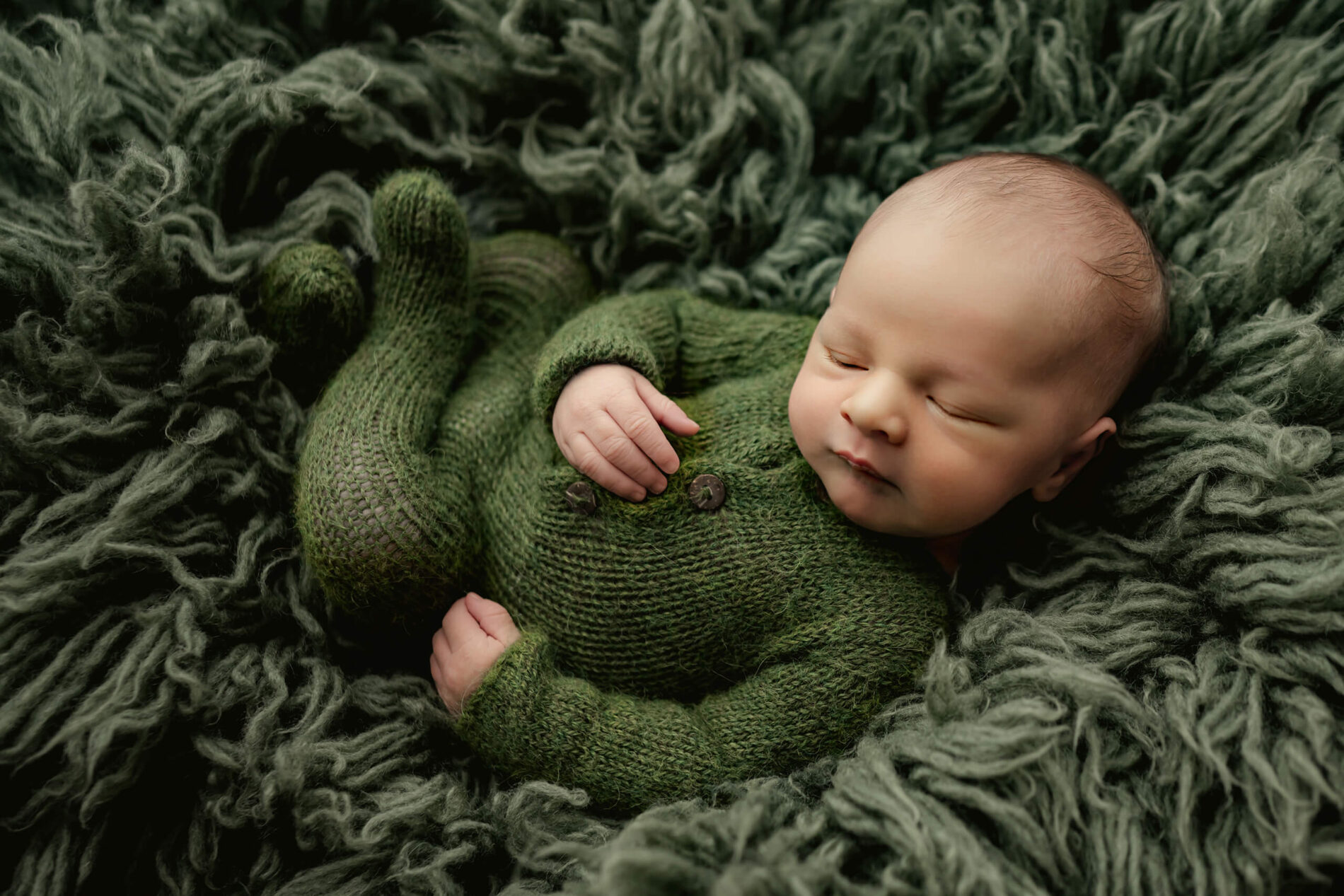 Newborn photography with props. Sleeping boy in knitted green onesie on top of a green cozy rug.