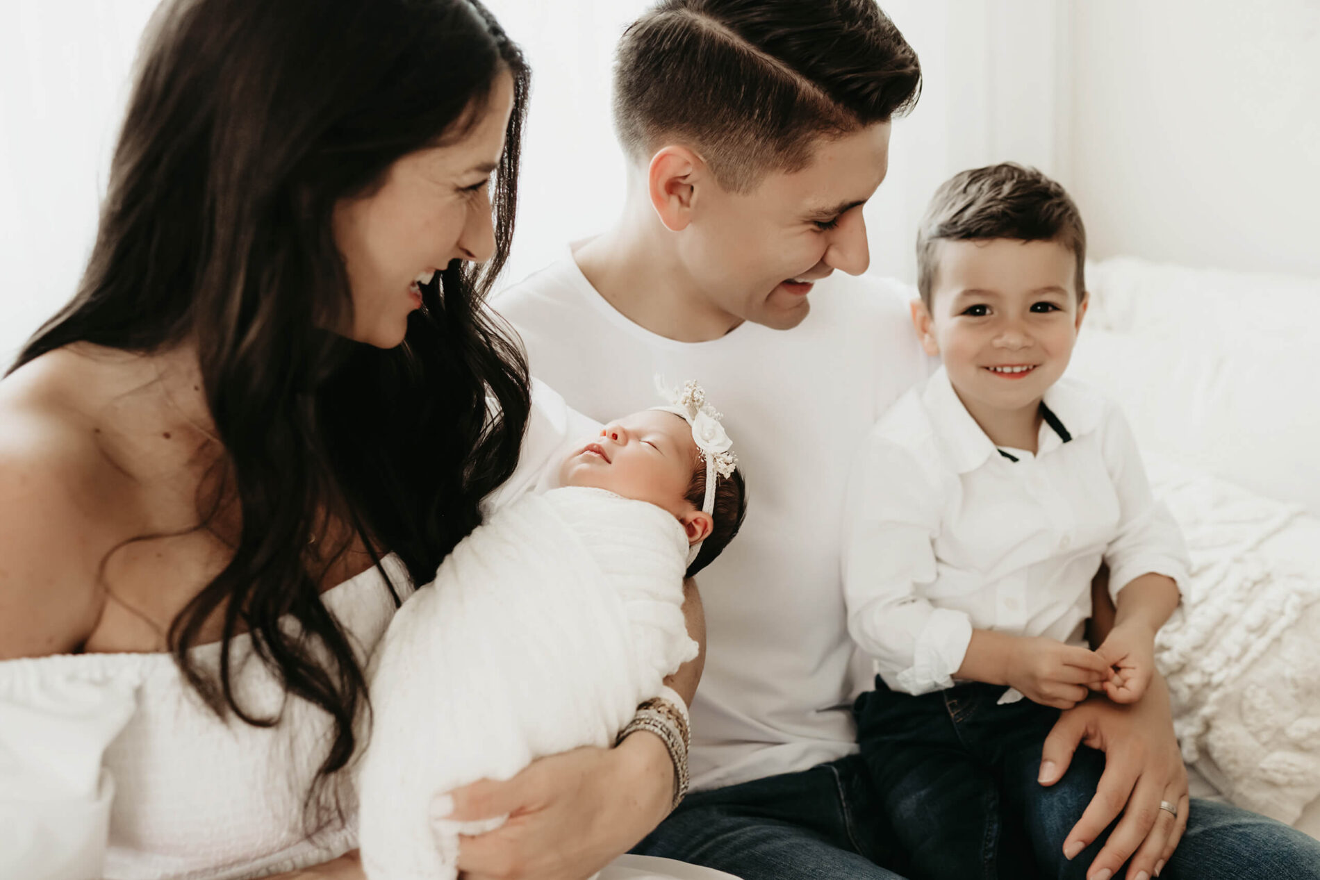 Natural family newborn photography in a Seattle studio. Smiling mom and dad sitting on a couch with toddler son and sleeping newborn daughter.