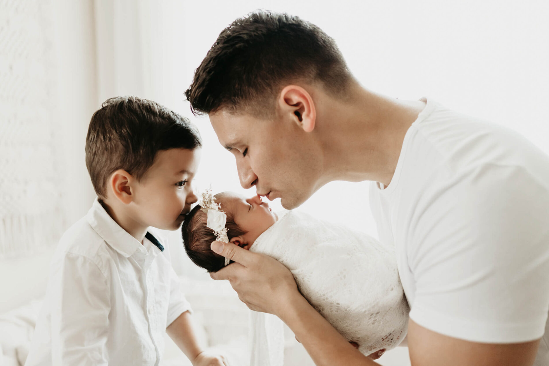 Unique newborn family pose. Dad holding his baby daughter and kissing her on her nose with her toddler brother kissing her on the head.