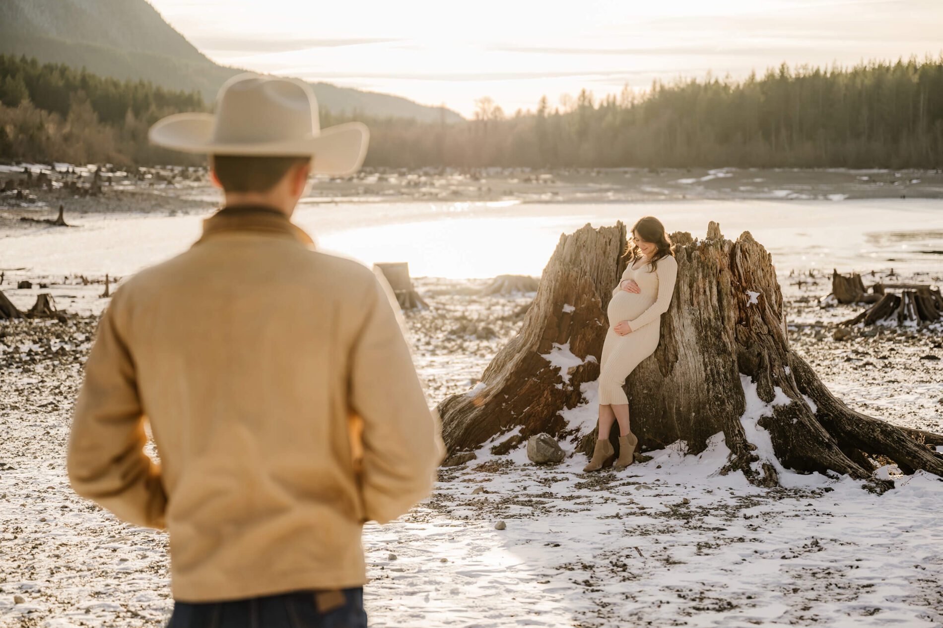 Seattle maternity photography with a young couple. Young pregnant woman leaning on a tree trunk looking at her belly. Her husband shot from the back, wearing a cowboy hat, looking at her.