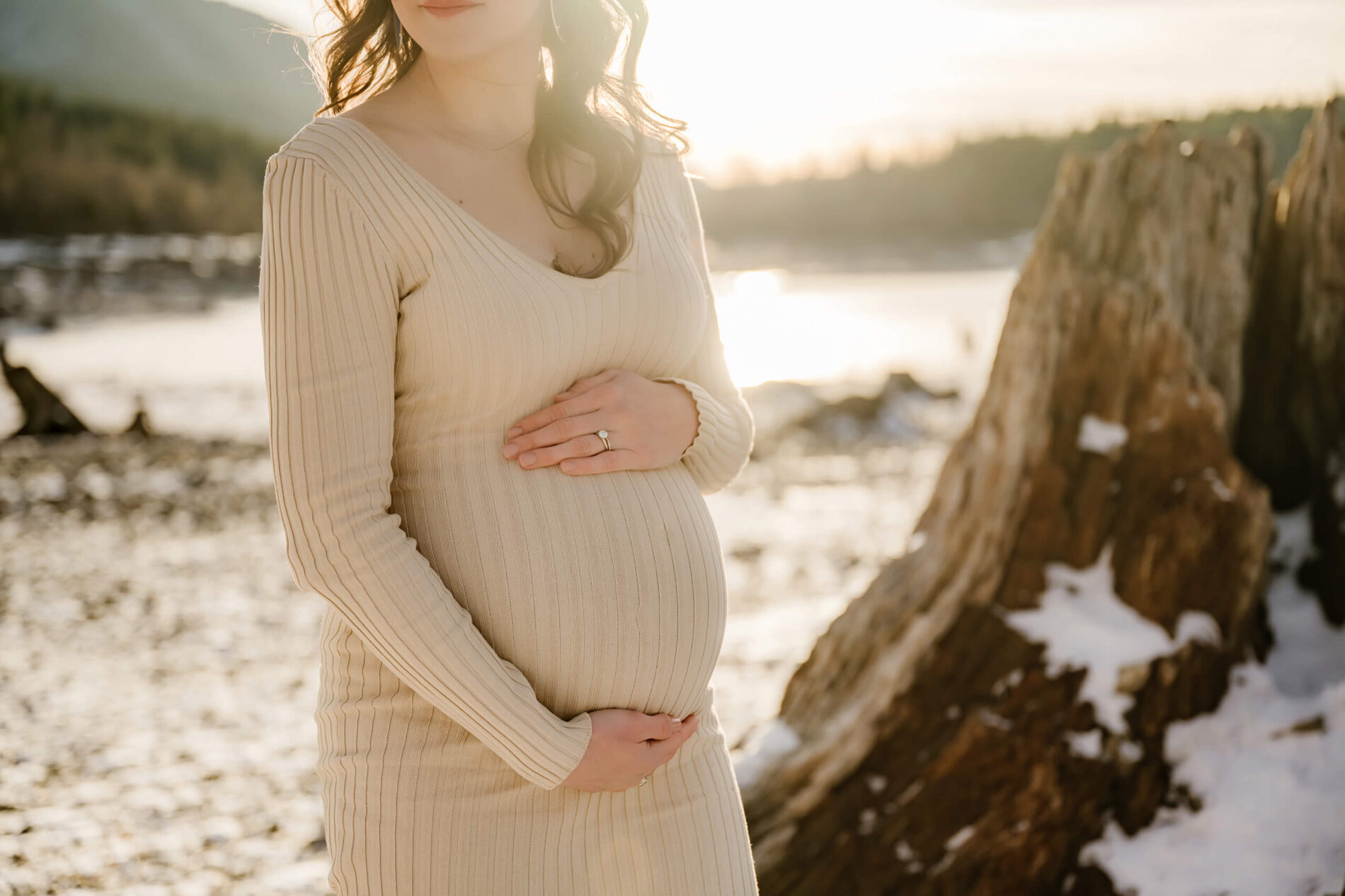 Seattle area maternity photo in the winter. Focus on pregnant woman wrapping her hands around her belly with frozen lake and mountains in the background