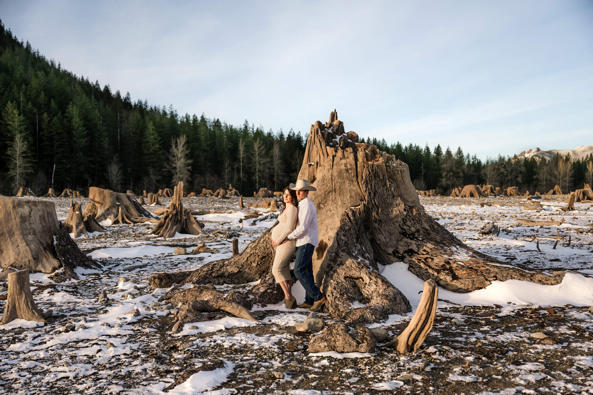A young couple posed in front of a large tree trunk during maternity photo session with beautiful PNW backdrop and snow on the ground