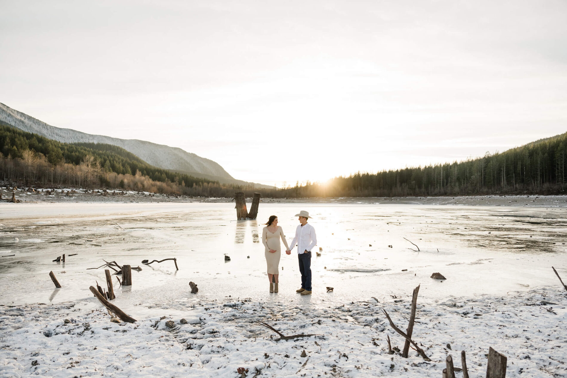 Couple posing during Seattle pregnancy photo shoot in front of a frozen lake at sunset. Beautiful mountains and trees in the background.