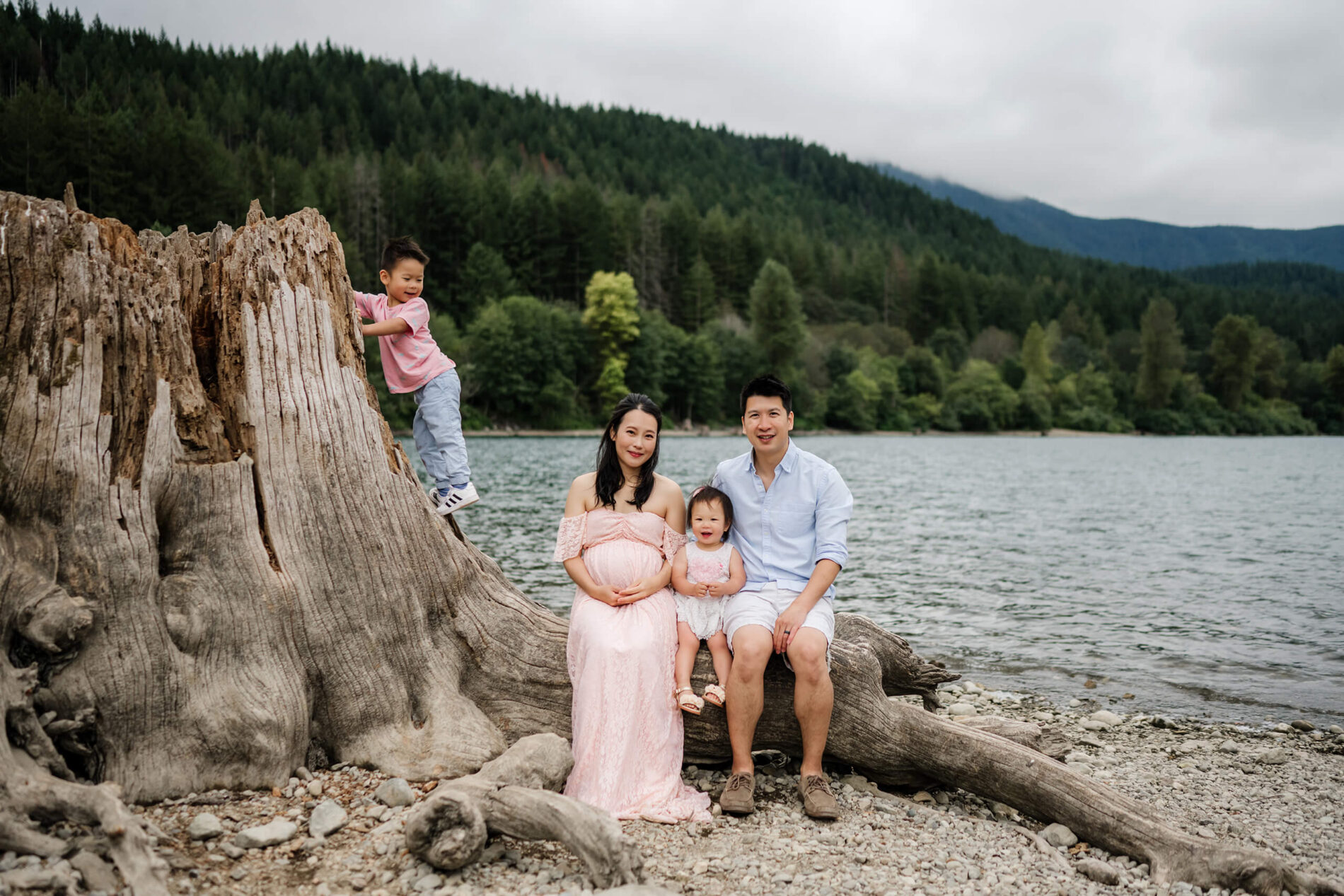 Family of four posed during a summer photo shoot on a large tree trunk with mountains and lake behind them