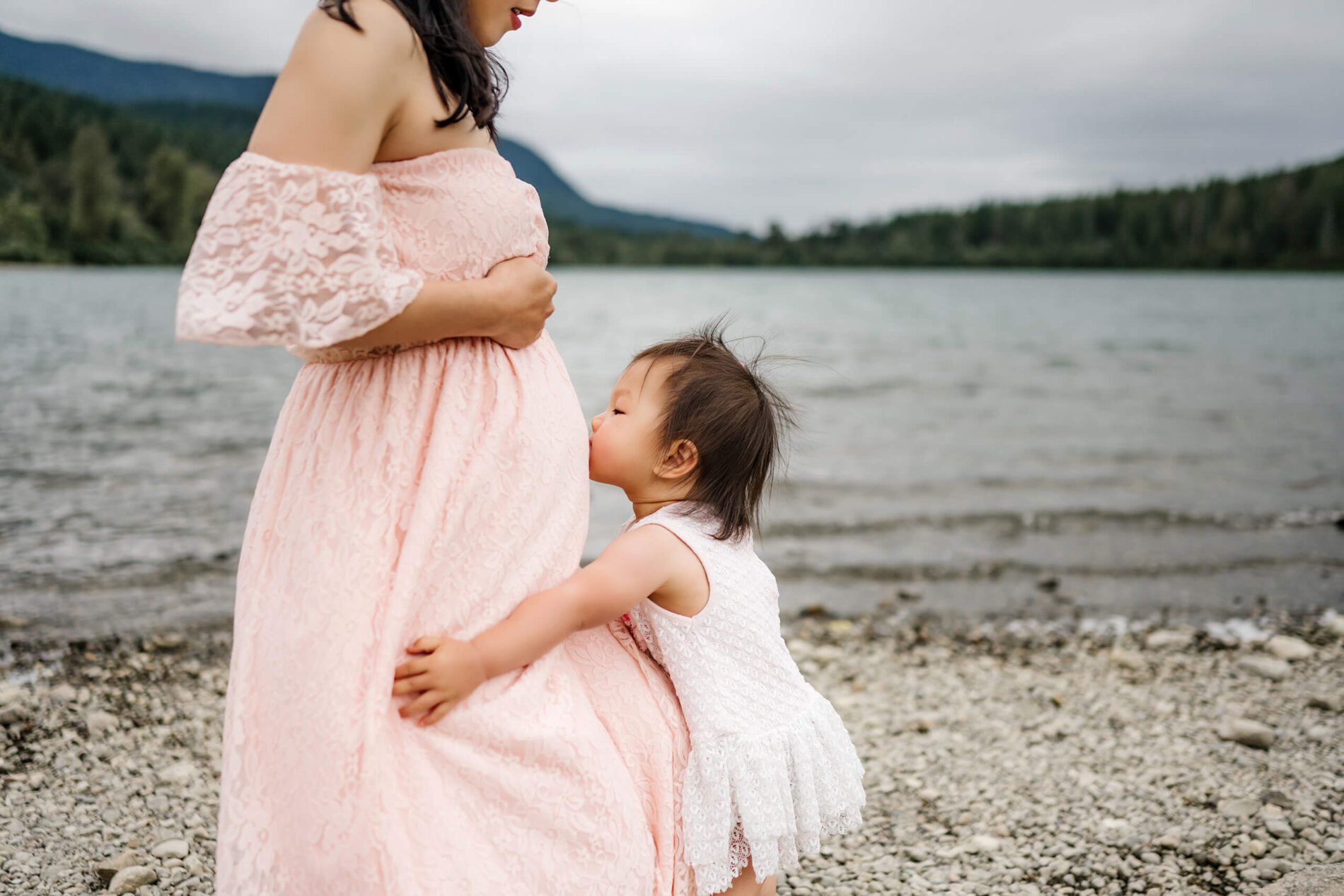 Daughter kissing mom's tummy. Mom and daughter are photographed on a beach in front of a lake with Cascades in the background