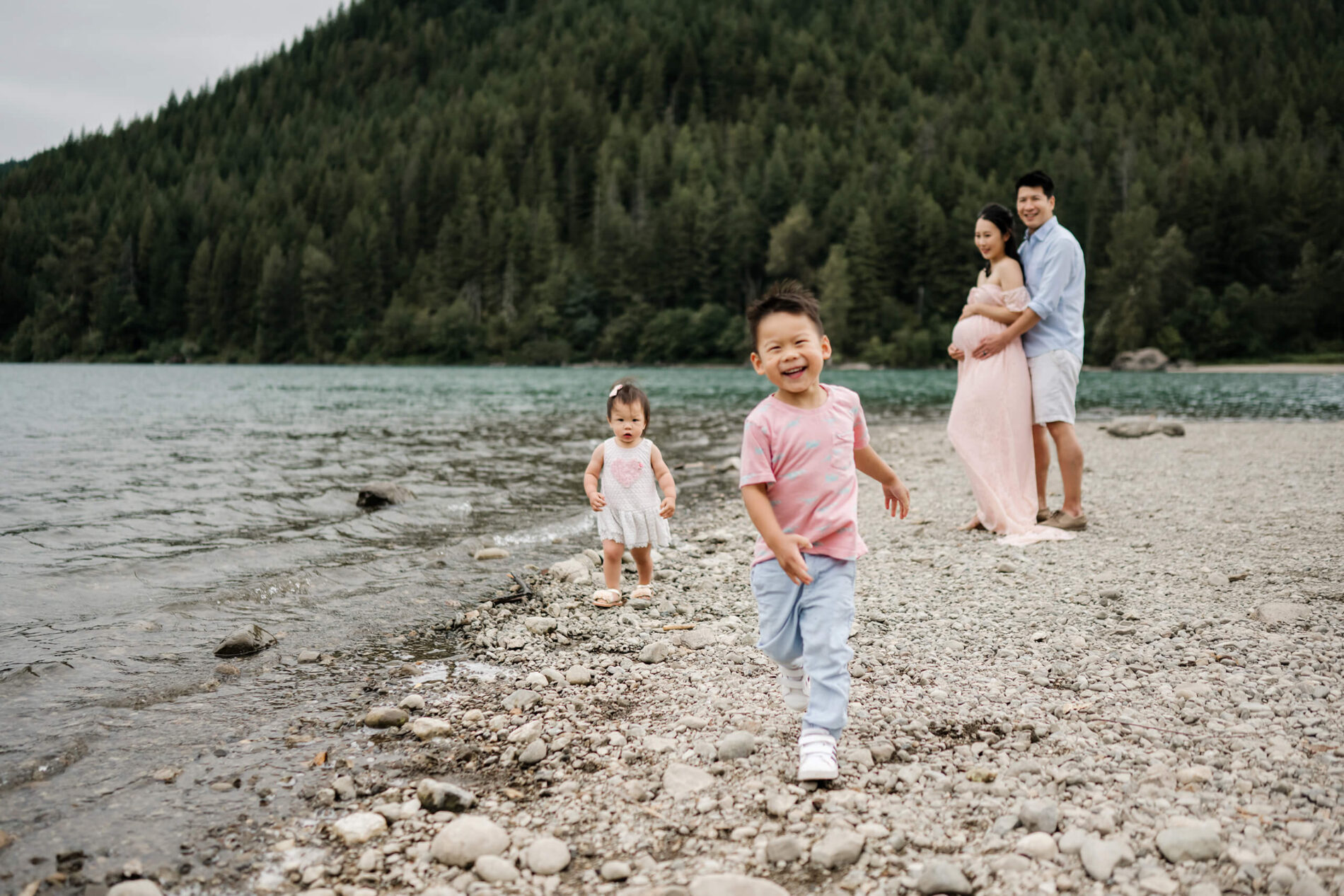 A lifestyle family photography session at Rattlesnake Lake. Dad hugging mom while their two children are running on the beach of a lake, with mountains in the background