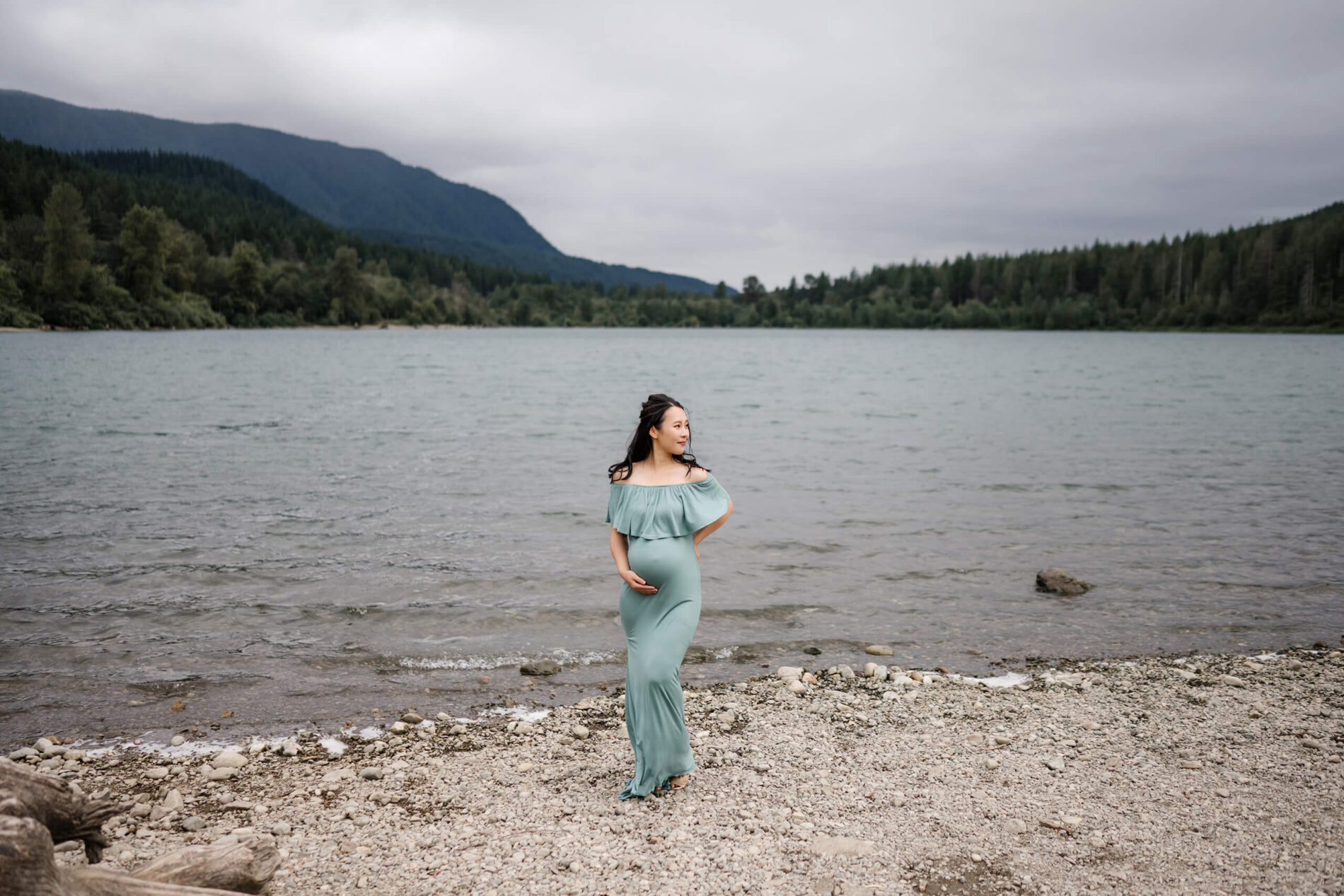 Pregnant woman in a green dress standing on a beach with lake and Cascades behind her.