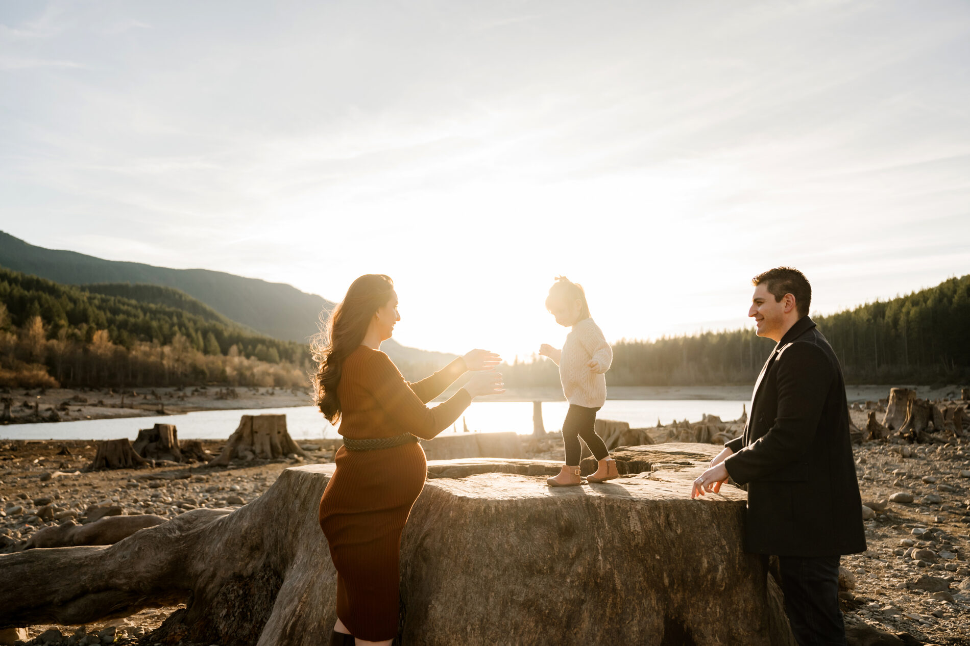 Lifestyle family photography with mountains, lake and trees in the background. Mom and dad playing with their daughter during a golden hour photo session in Seattle.