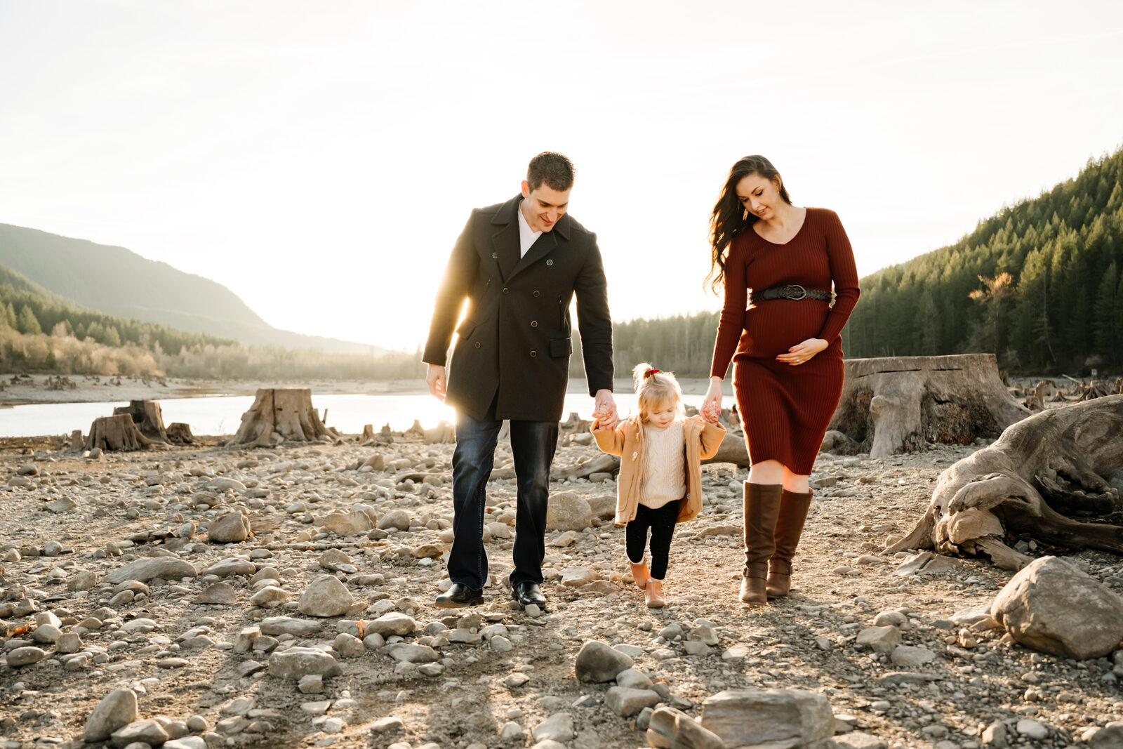 Winter family photography in Seattle area. Cozily dressed mom and dad walking with their daughter on a beach with lake and mountains in the background.