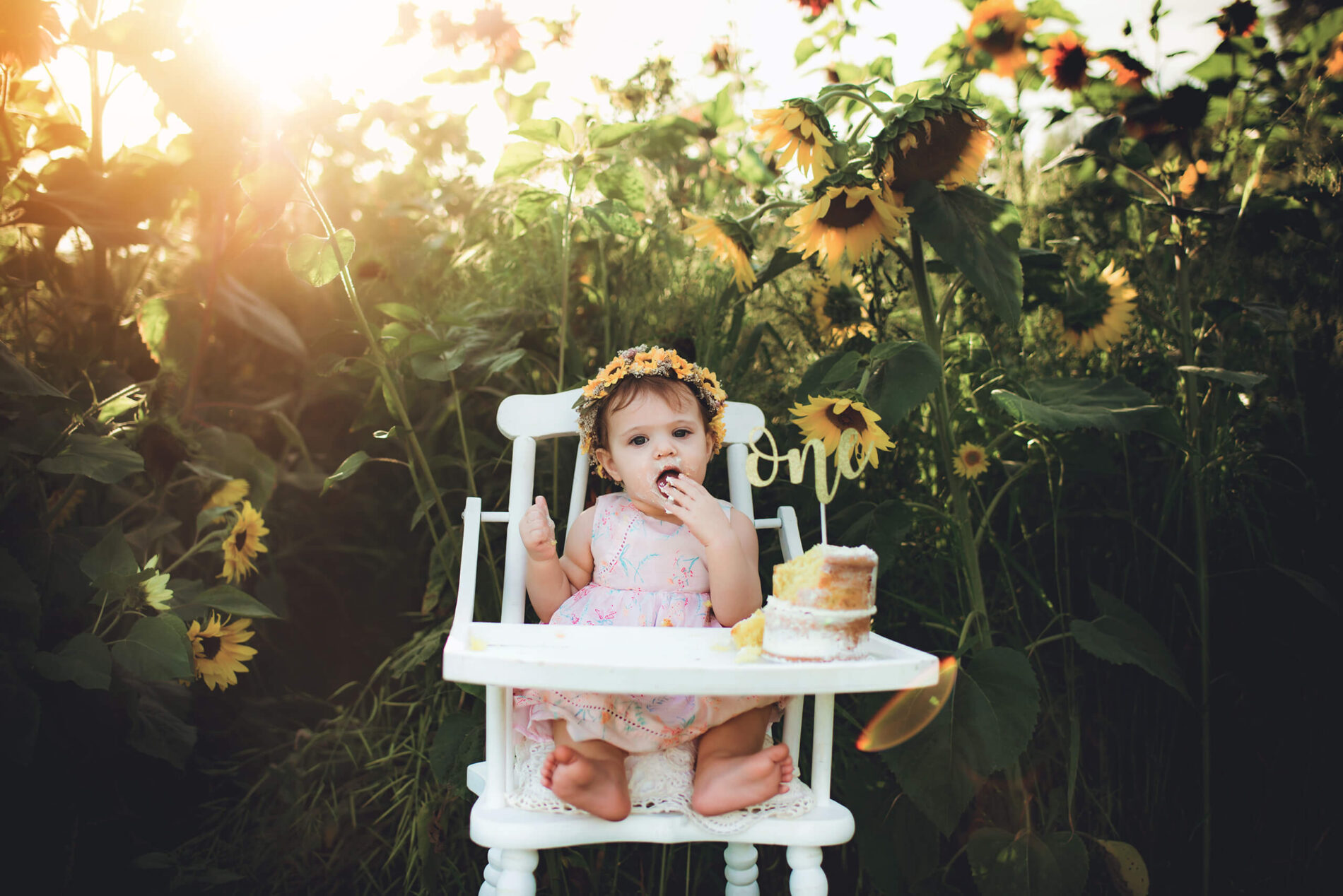 One year old girl in a white high chair eating cake with her hands during cake smash photo session