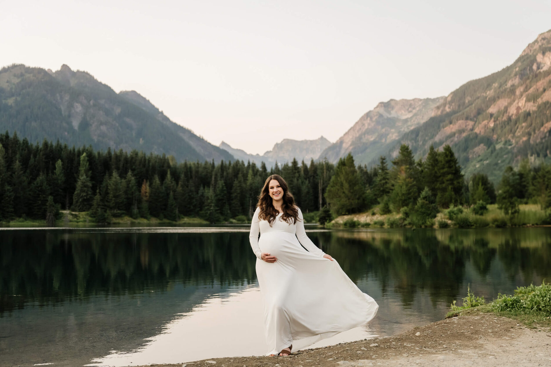 Pregnant woman in a white dress posing by the lake with mountains in the background