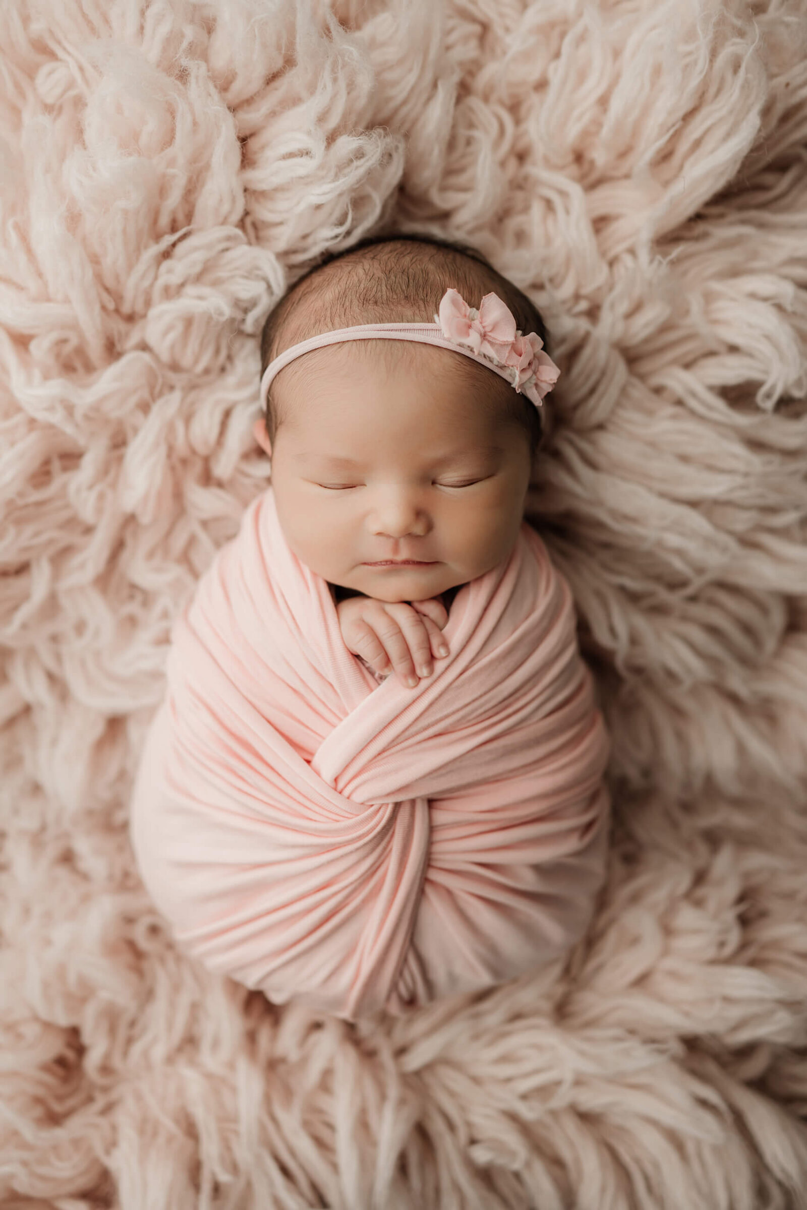 Sleeping newborn baby girl wrapped in pink and wearing a pink floral headband on a textured pink blanket.