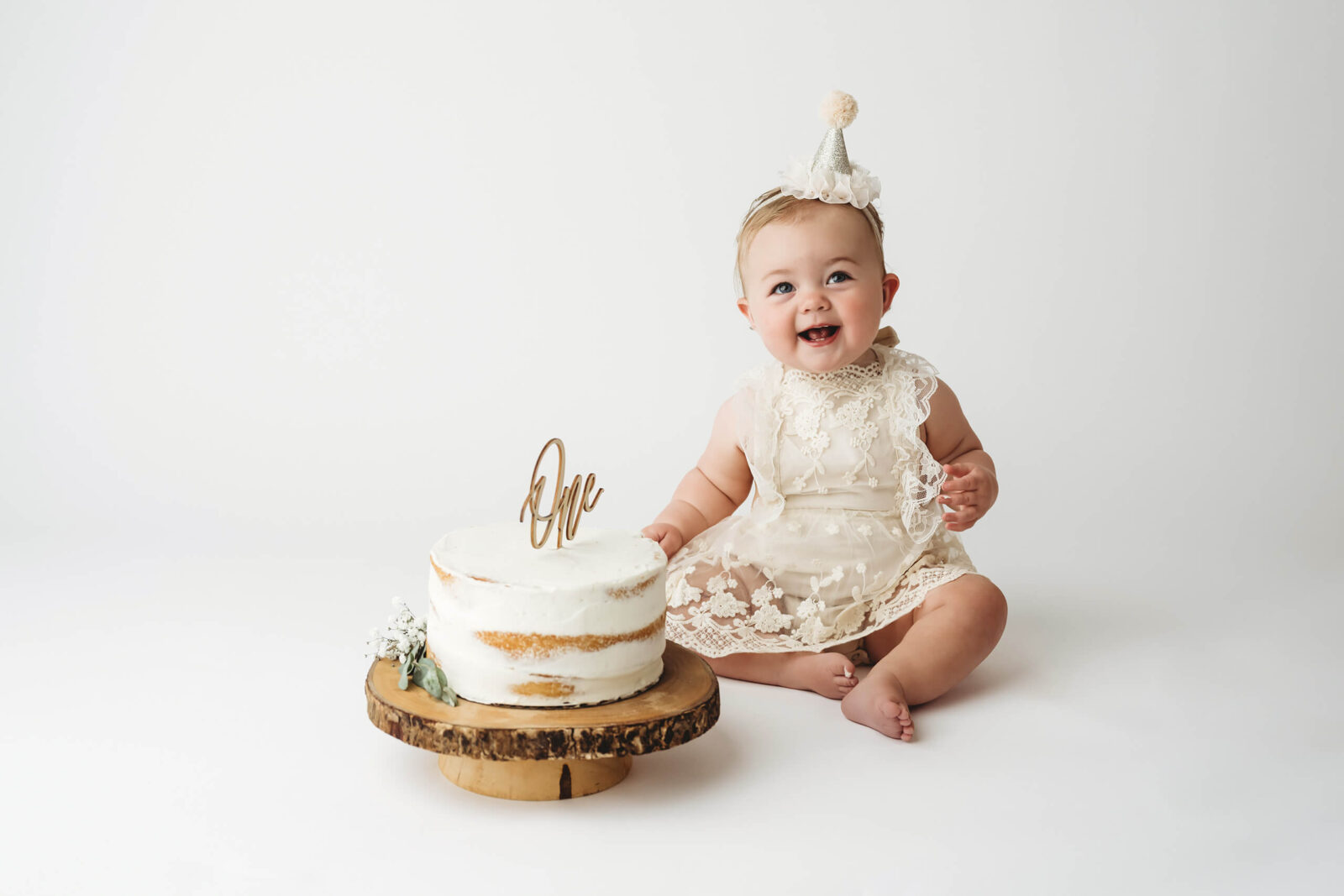 Seattle first-birthday milestone photo: smiling baby girl in lace dress and party hat sits beside rustic naked cake with “one” topper on white backdrop—Kirkland studio by Lana Sky Photography. 