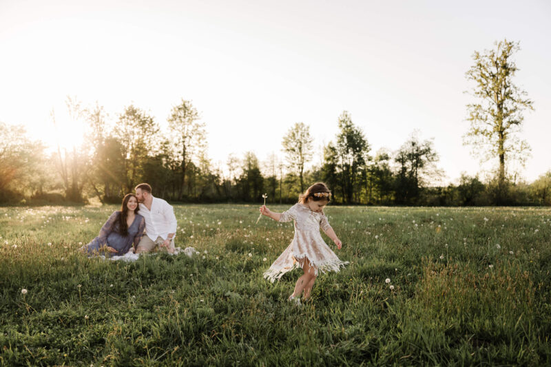 Golden-hour Seattle maternity photo: pregnant mom in lavender dress on blanket as husband kisses her forehead; daughter twirls with dandelion in grassy Eastside meadow — Lana Sky Photography.