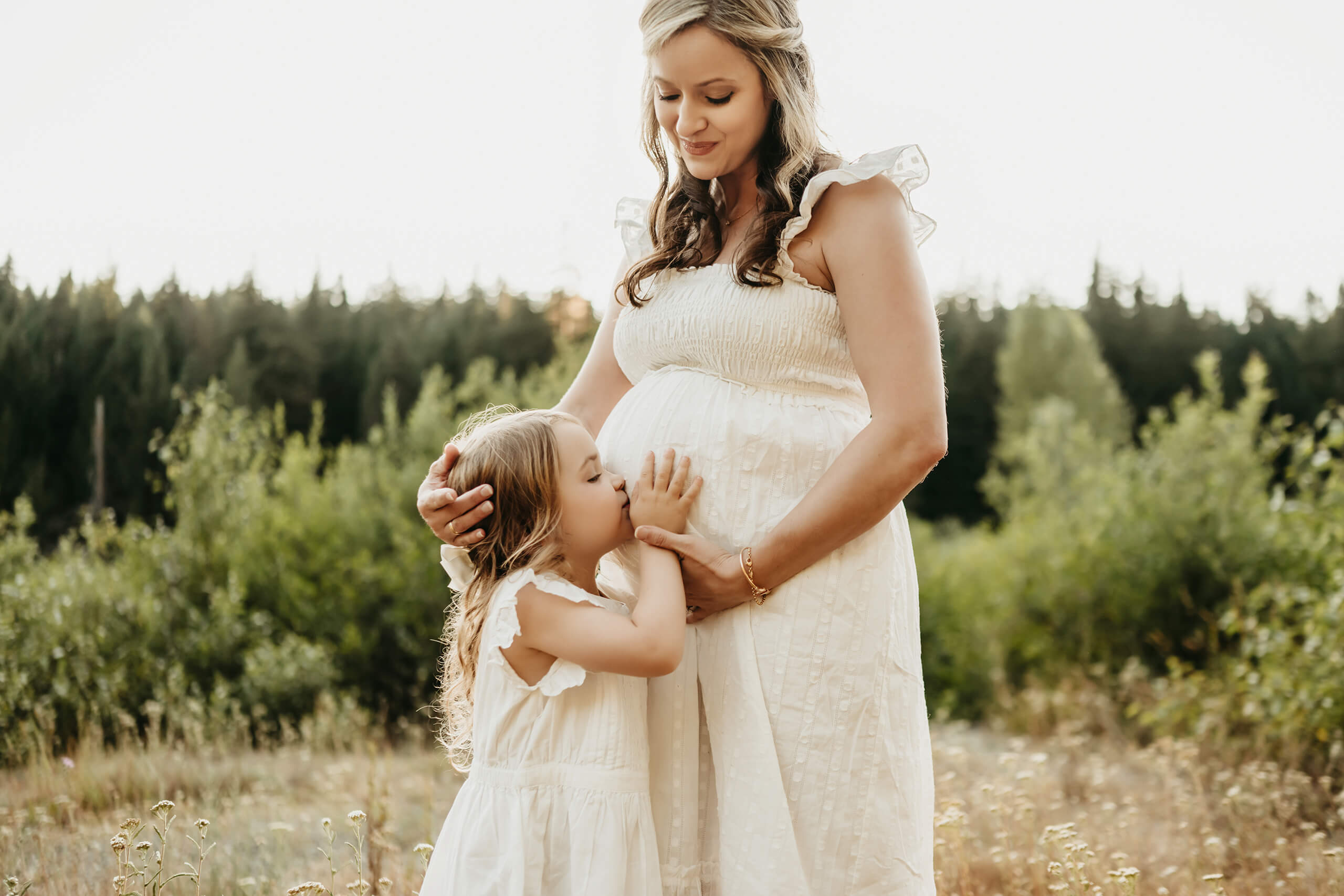 Maternity photoshoot in Seattle area: little girl in white sundress kisses her pregnant mom’s belly during golden-hour meadow session framed by evergreen forest