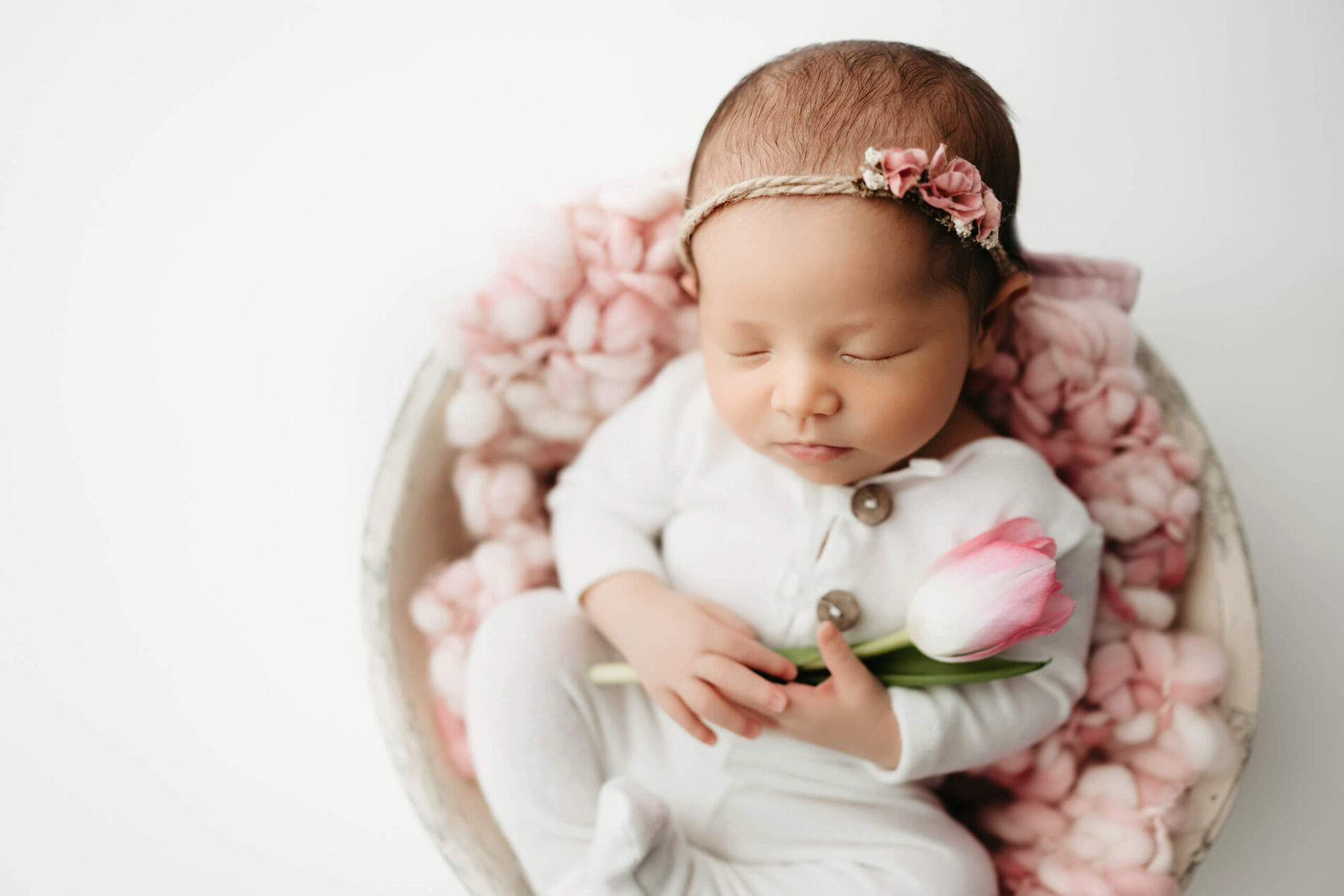 Sleeping newborn girl in a white romper, fingers wrapped around a single blush tulip, nestled in a bowl lined with pink wool.