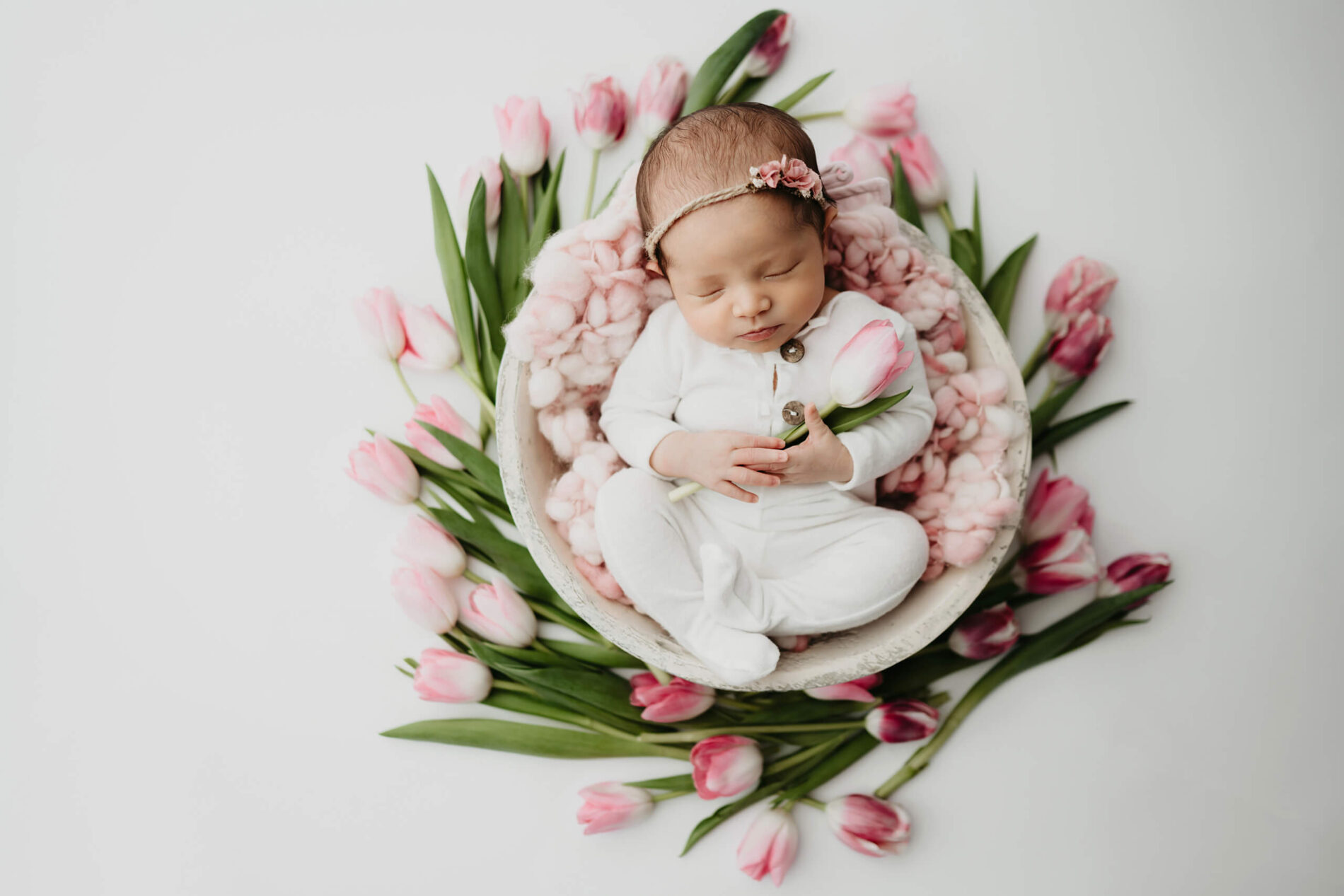 Newborn sleeping in bowl with blush tulips tucked around, soft studio light.