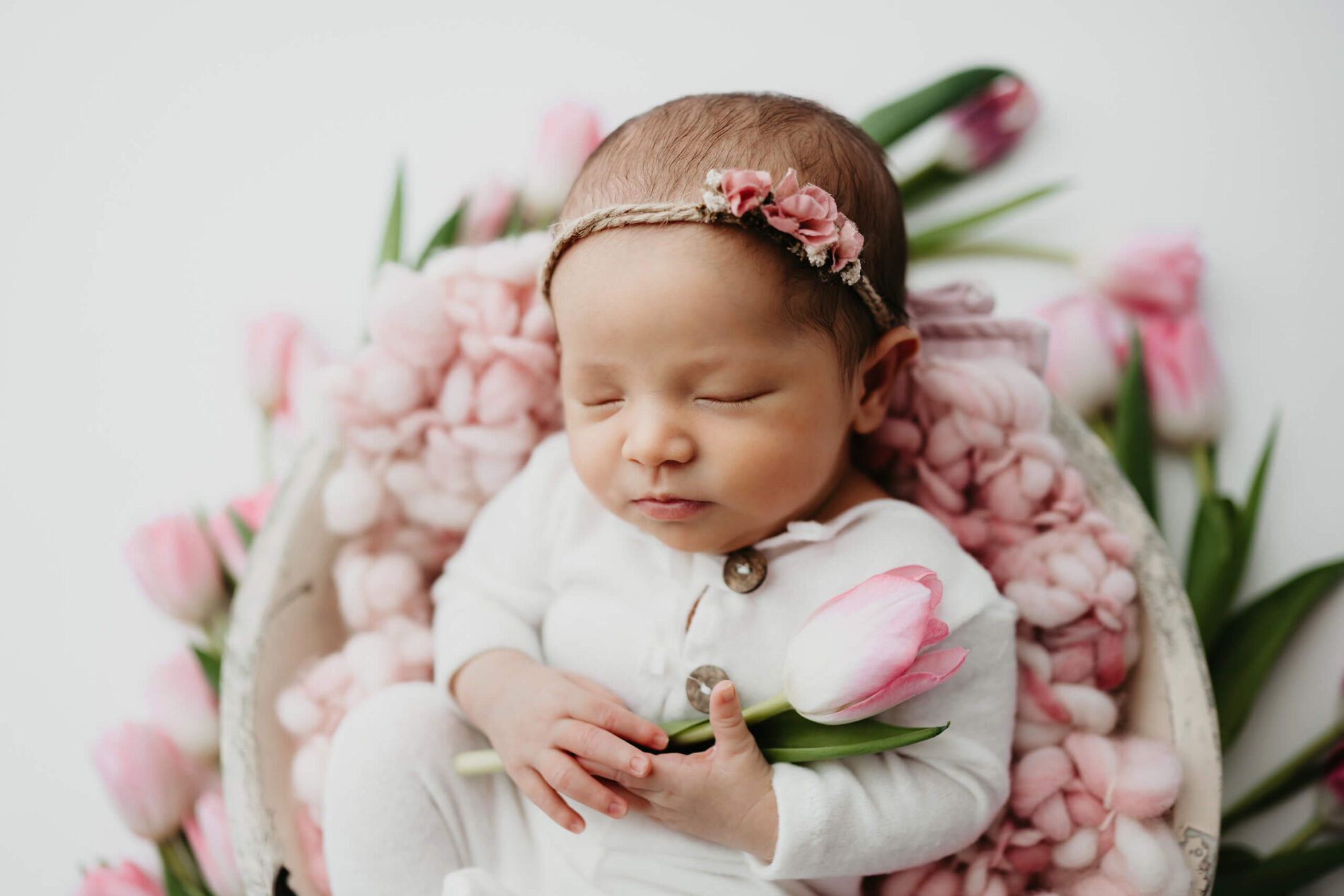 Sleeping infant clutching a blush tulip, nestled on soft pink wool inside a round prop bowl.