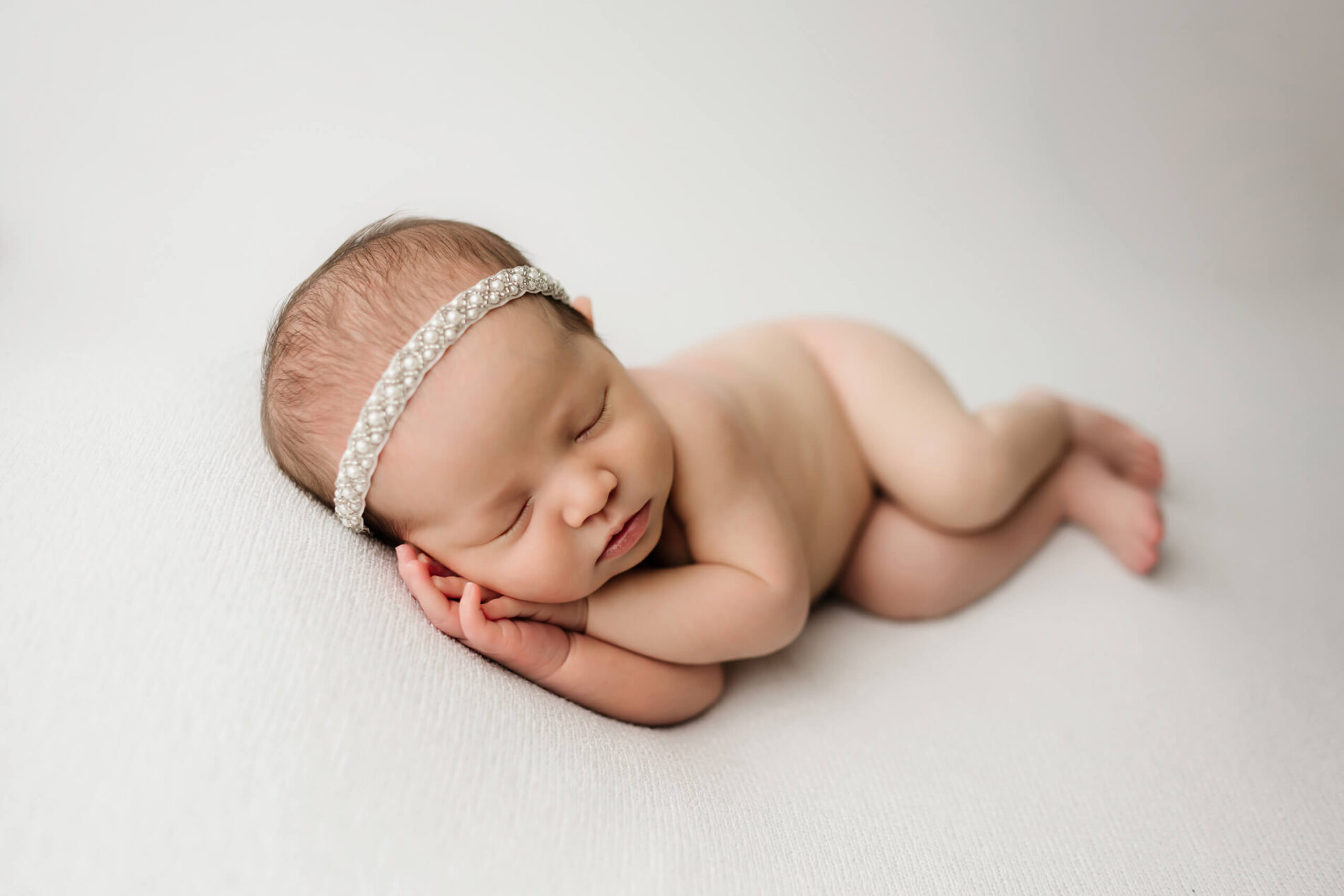Sleeping newborn resting on cream blanket, pearl headband catching the soft studio light.