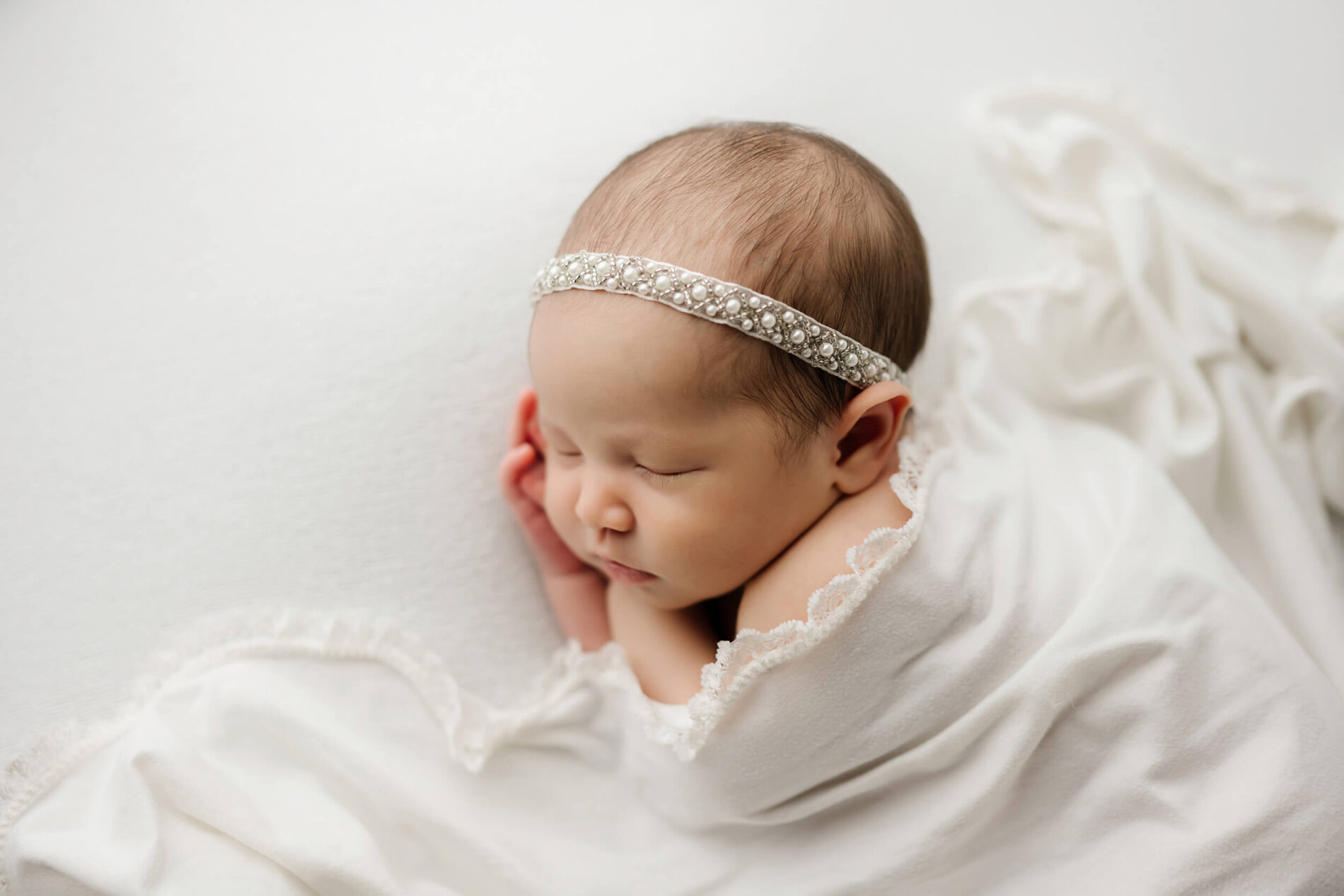 Seattle newborn photography: sleeping baby swaddled in white, pearl headband, on creamy backdrop, captured in Kirkland studio by Lana Sky Photography.