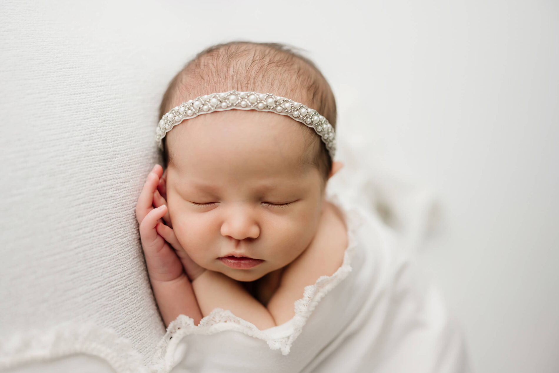 Close-up of newborn wrapped in ivory swaddle, peaceful face and pearl headband