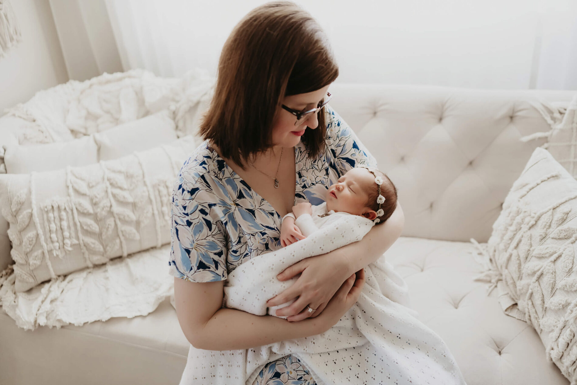 Soft neutral couch scene—mom cradles baby in eyelet blanket, gazing down with a gentle smile.