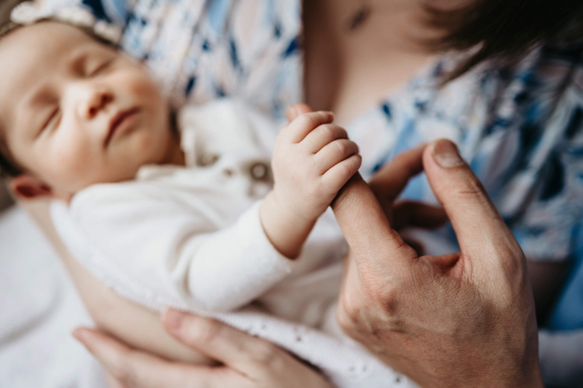 Macro moment: tiny newborn fingers curl around dad’s index finger while she sleeps peacefully in mom’s arms.