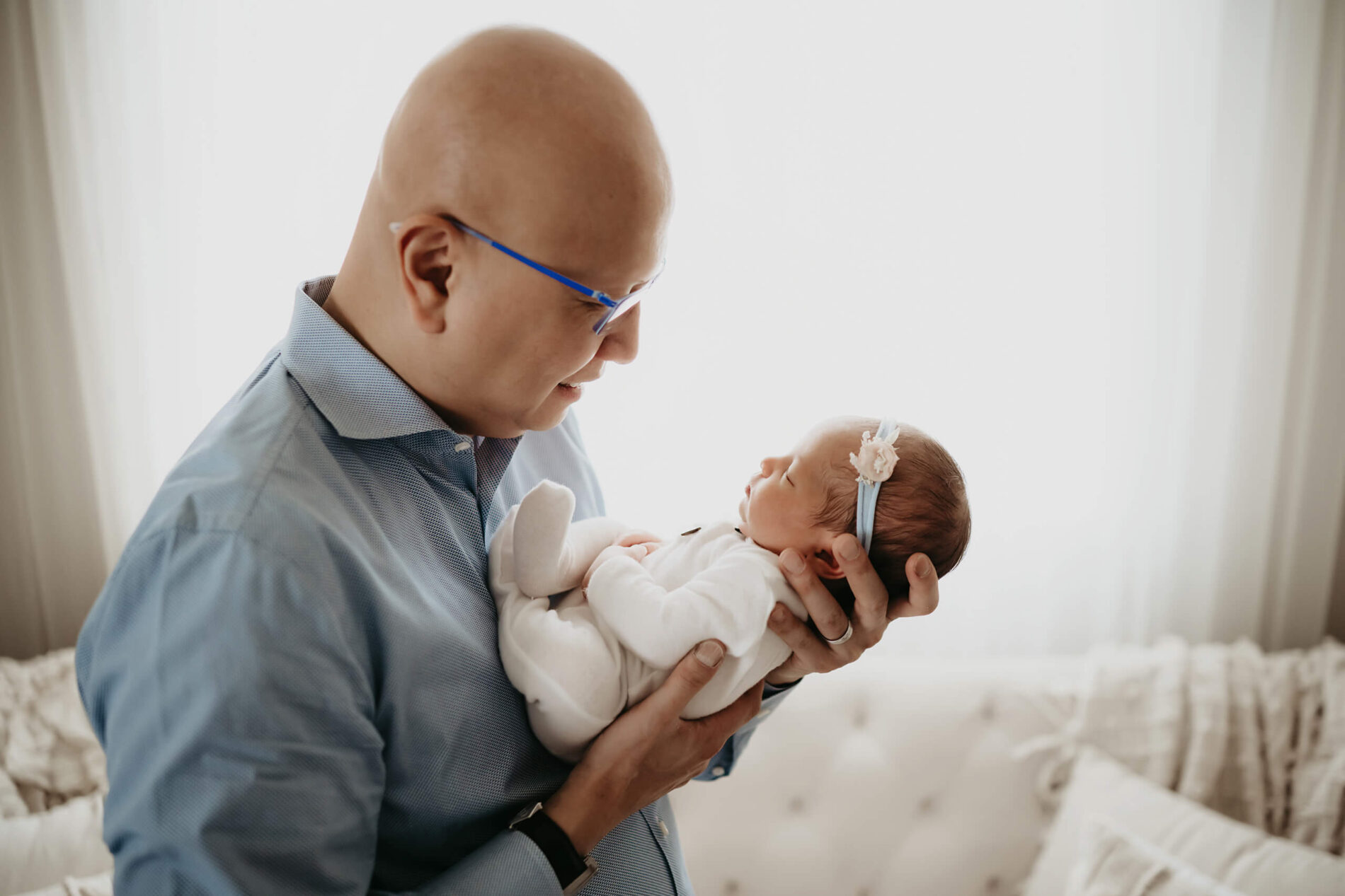 Dad lifts his baby girl toward the window light, blue glasses and shirt echoing her pastel headband.