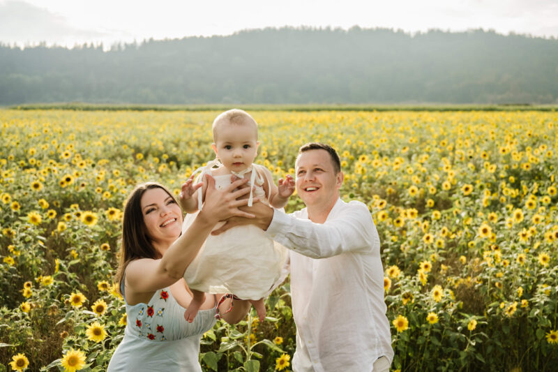 Young parents lift their smiling baby girl above shoulder-height in a vast sunflower field near Seattle, glowing with late-afternoon golden light and a tree-lined hill in the distance.