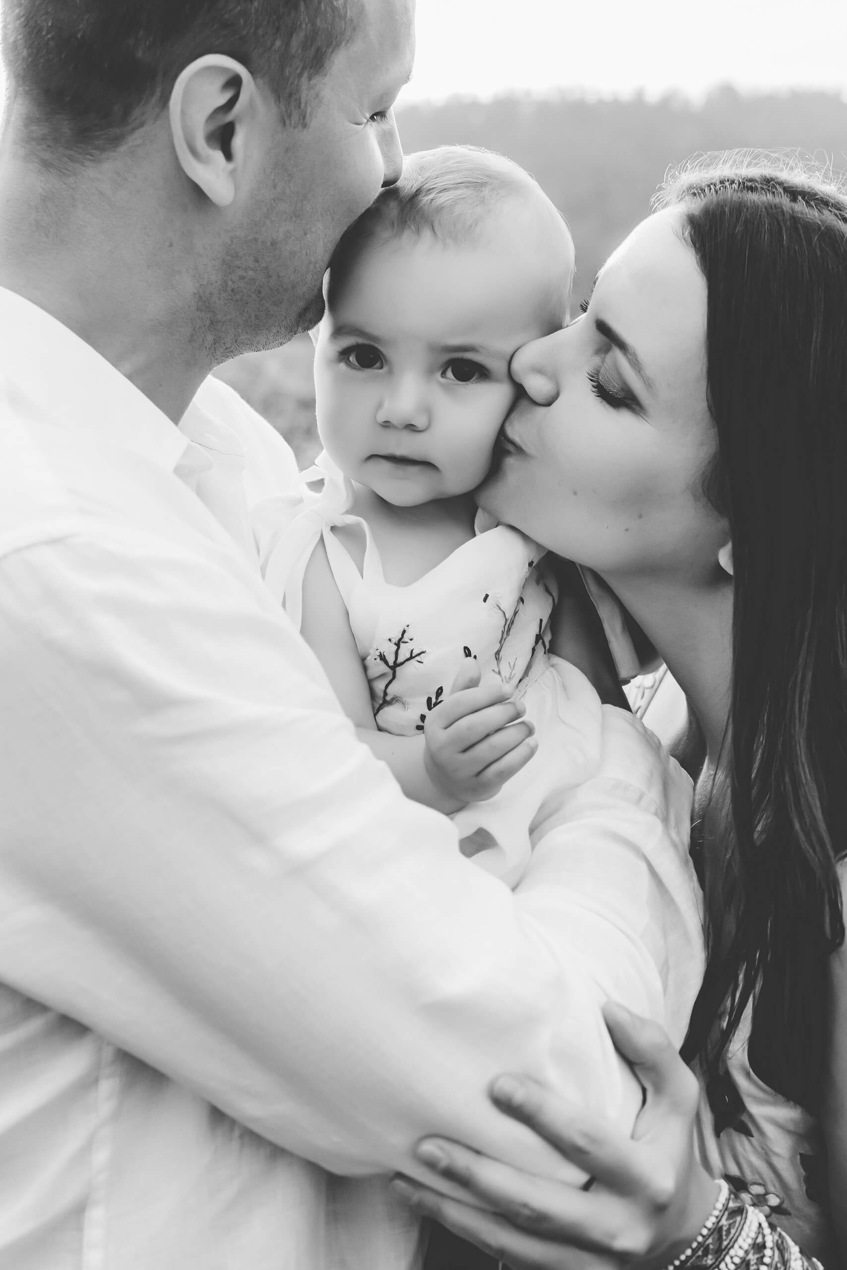 Close black-and-white shot of parents kissing their baby’s head and cheek as the baby gazes calmly at the camera.