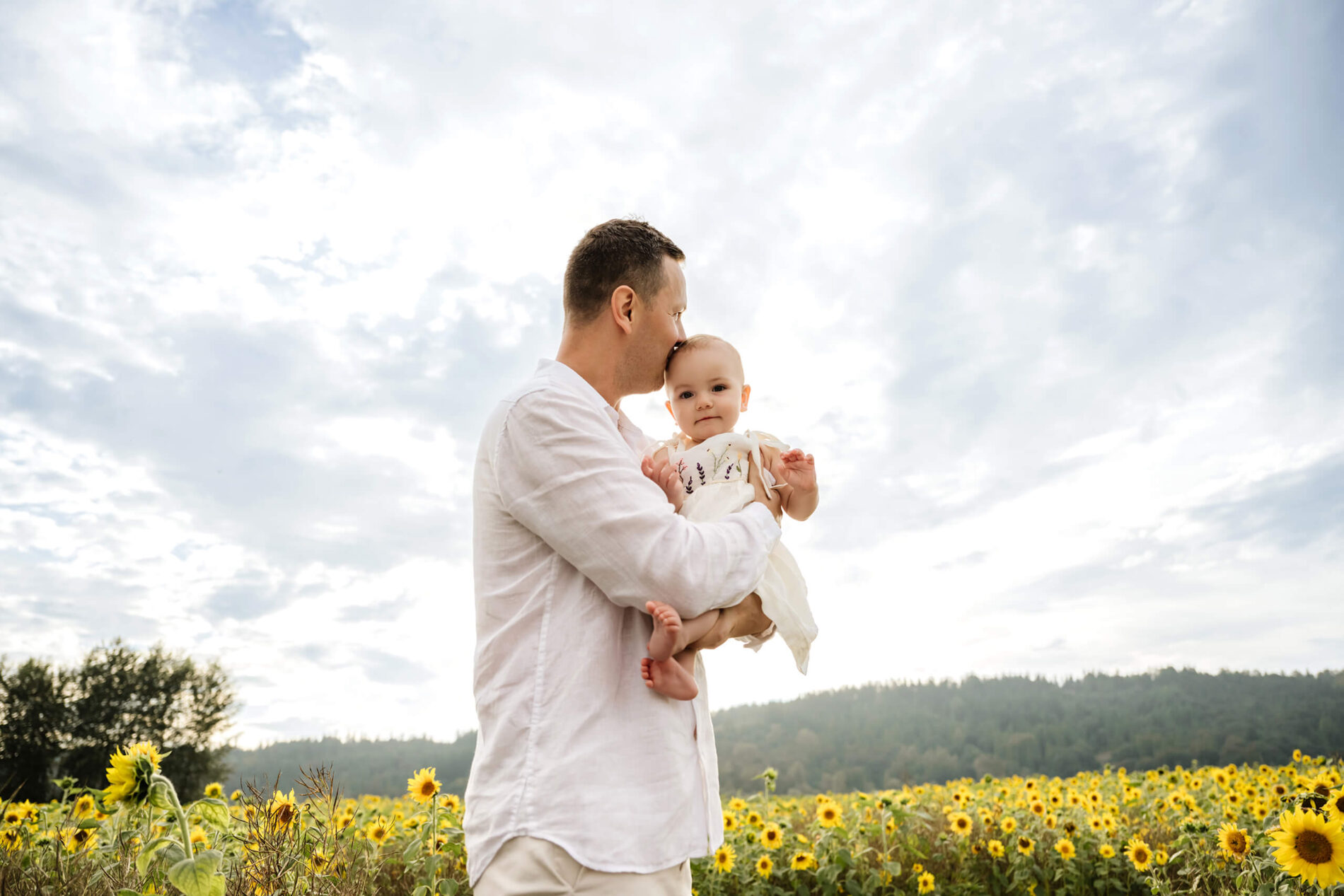 Father cradles his baby against a dramatic sky, kissing her head above a field of sunflowers.